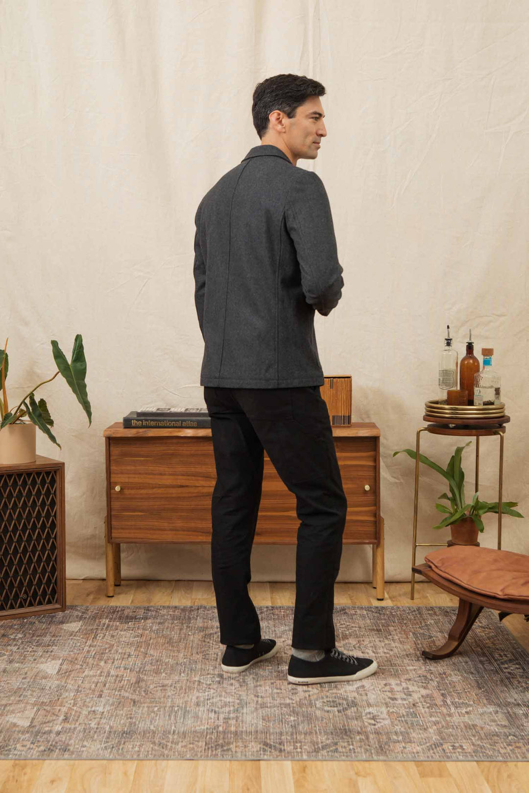A man faces away in a room with a neutral rug, mid-century modern furniture, plants, and bottles. He wears the Bridge & Burn Nate Chore Blazer in Onyx Wool with black pants and sneakers.