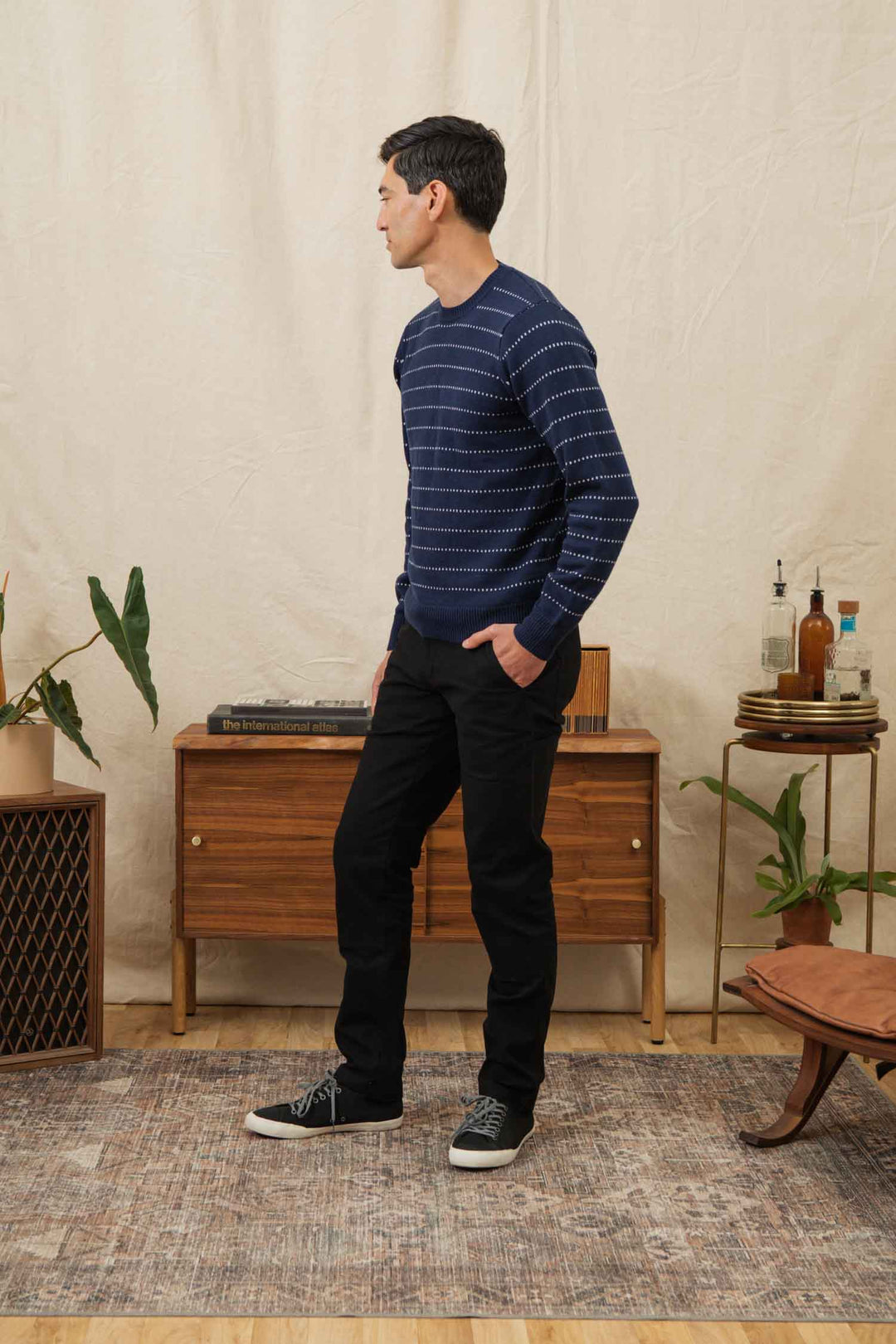 A man wearing the Bridge & Burn Orson Sweater in Navy Dotted Path, black pants, and black sneakers stands sideways in a living room with mid-century modern furniture, plants, and a beige backdrop.