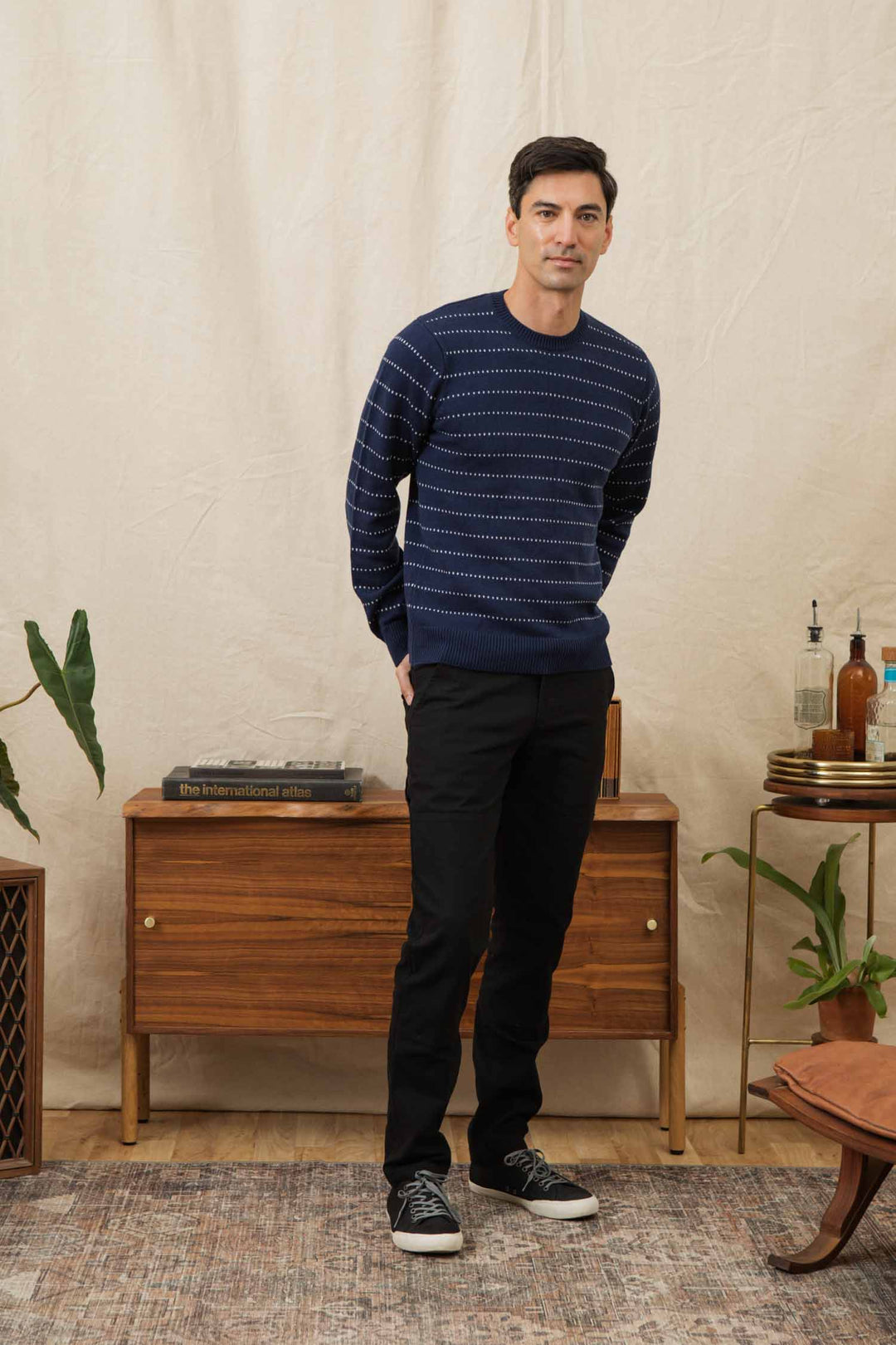 A man stands indoors in front of a wooden sideboard, wearing the Bridge & Burn Orson Sweater in Navy Dotted Path, black pants, and black sneakers. Decorative plants and bottles are on the sideboard against a beige fabric curtain.