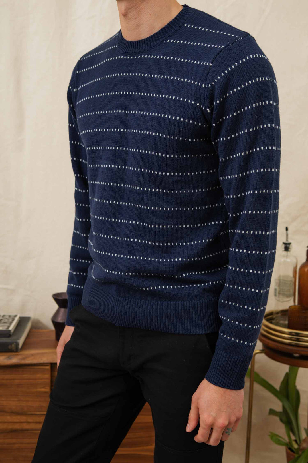 A person stands indoors near a wooden table with decor, wearing Bridge & Burn's Orson Sweater in Navy Dotted Path and black pants.