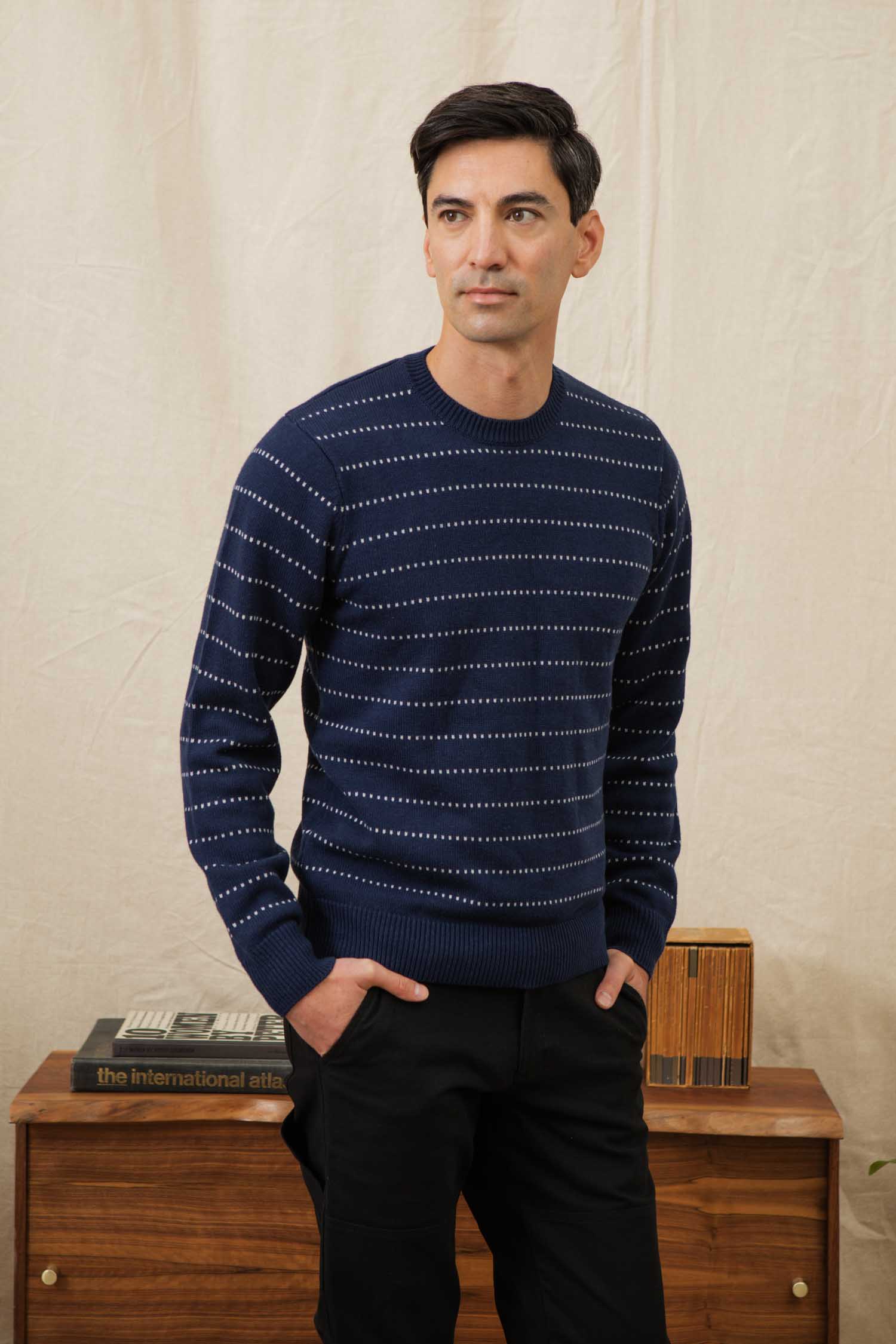 A man with short dark hair wears the Bridge & Burn Orson Sweater in Navy Dotted Path, standing with hands in pockets before a wooden dresser topped with books and decor, set against a beige background.