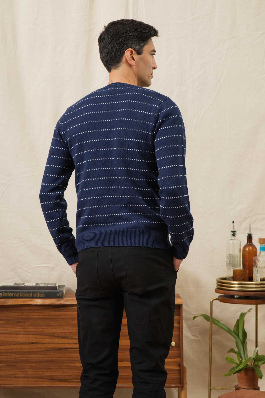 A man stands indoors near a wooden sideboard, wearing the Bridge & Burn Orson Sweater in Navy Dotted Path— a lightweight merino wool knit with white dotted stripes—paired with black pants; a plant is visible in the corner.