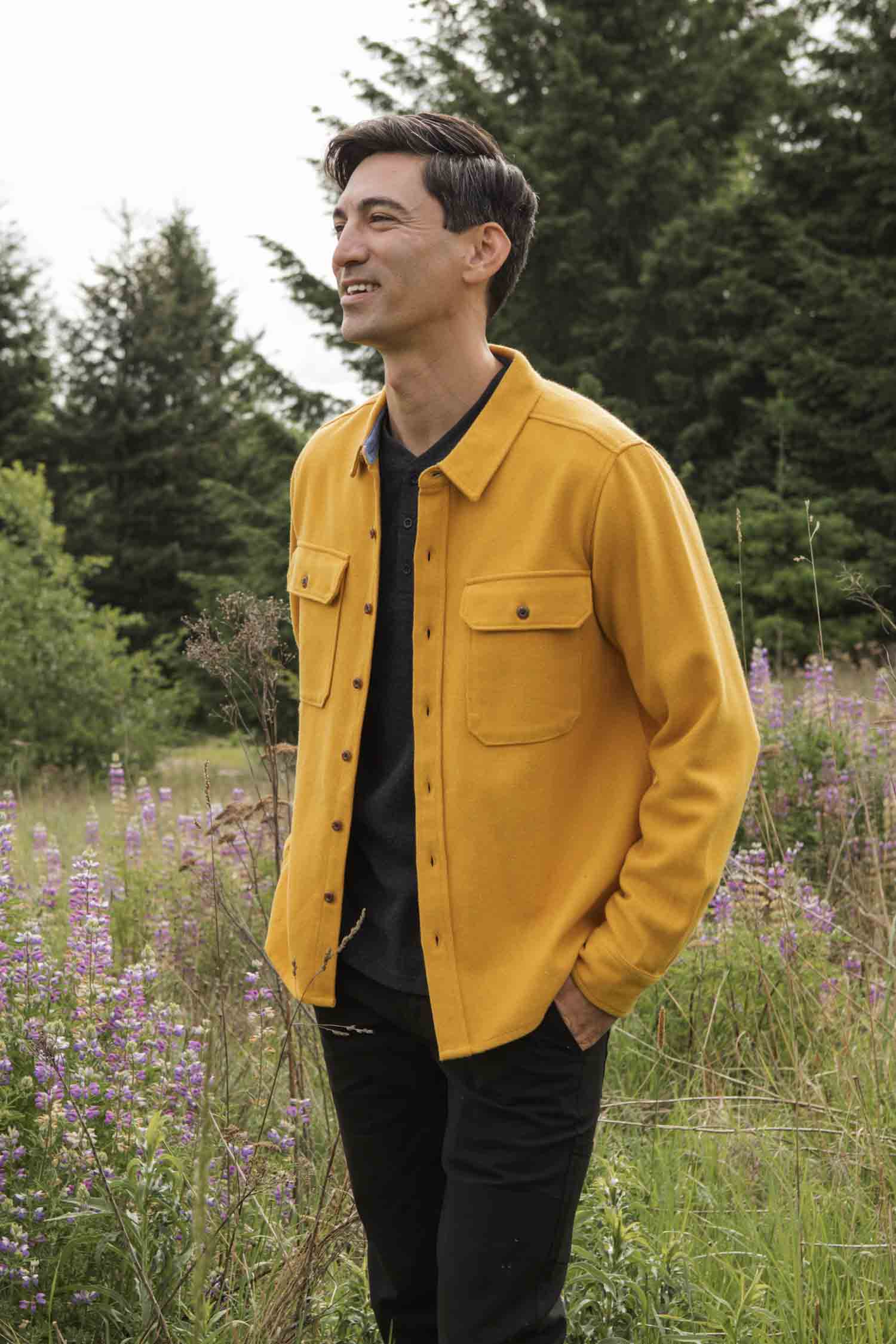 A man smiles outdoors in a grassy field wearing the Bridge & Burn Pointer Overshirt in Antique Gold and black pants, surrounded by purple wildflowers and green trees—showcasing sustainable style.