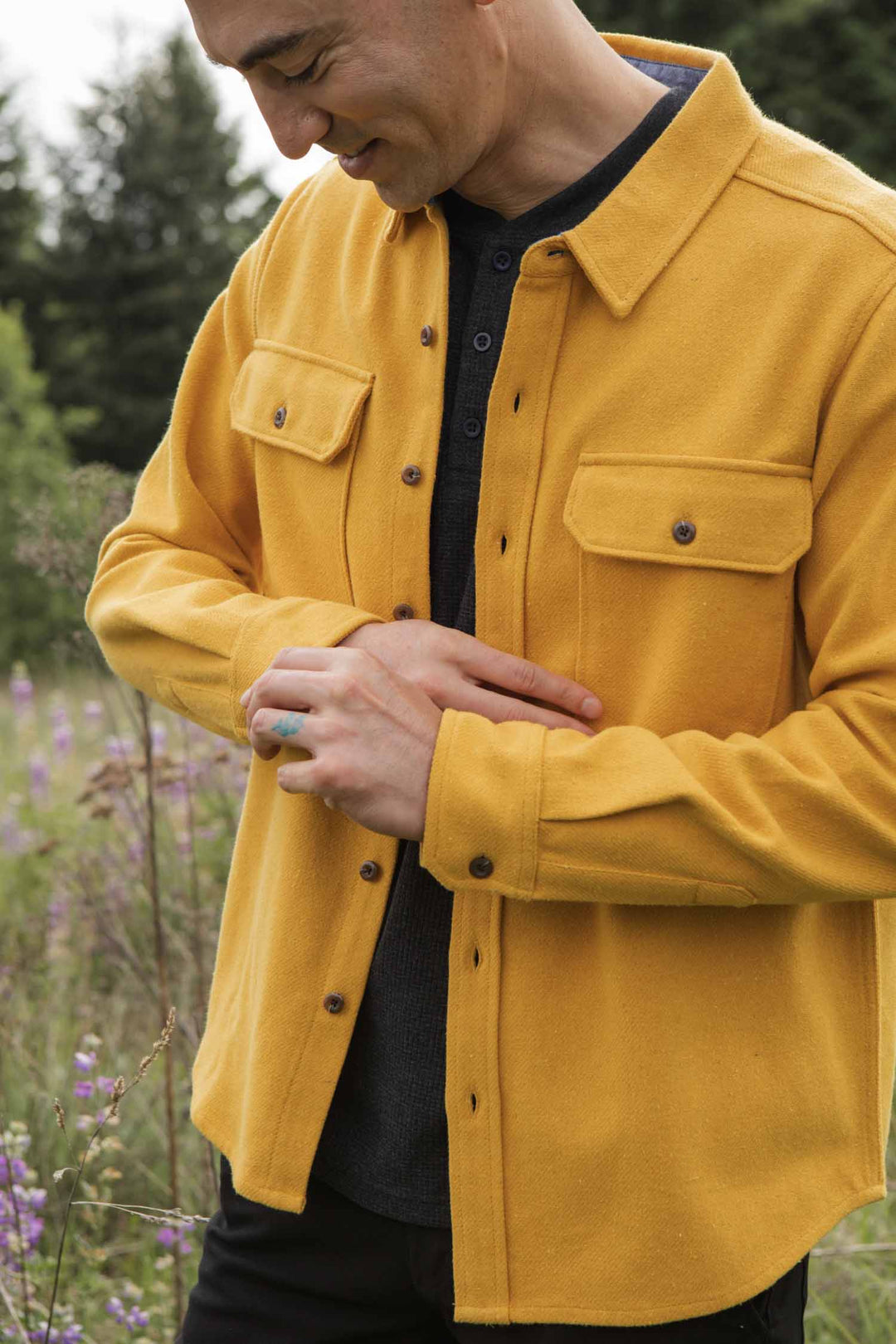 A person stands in a field, smiling and looking down with hands clasped, wearing the Bridge & Burn Pointer Overshirt in Antique Gold over a dark shirt. Trees and purple wildflowers create a peaceful backdrop.