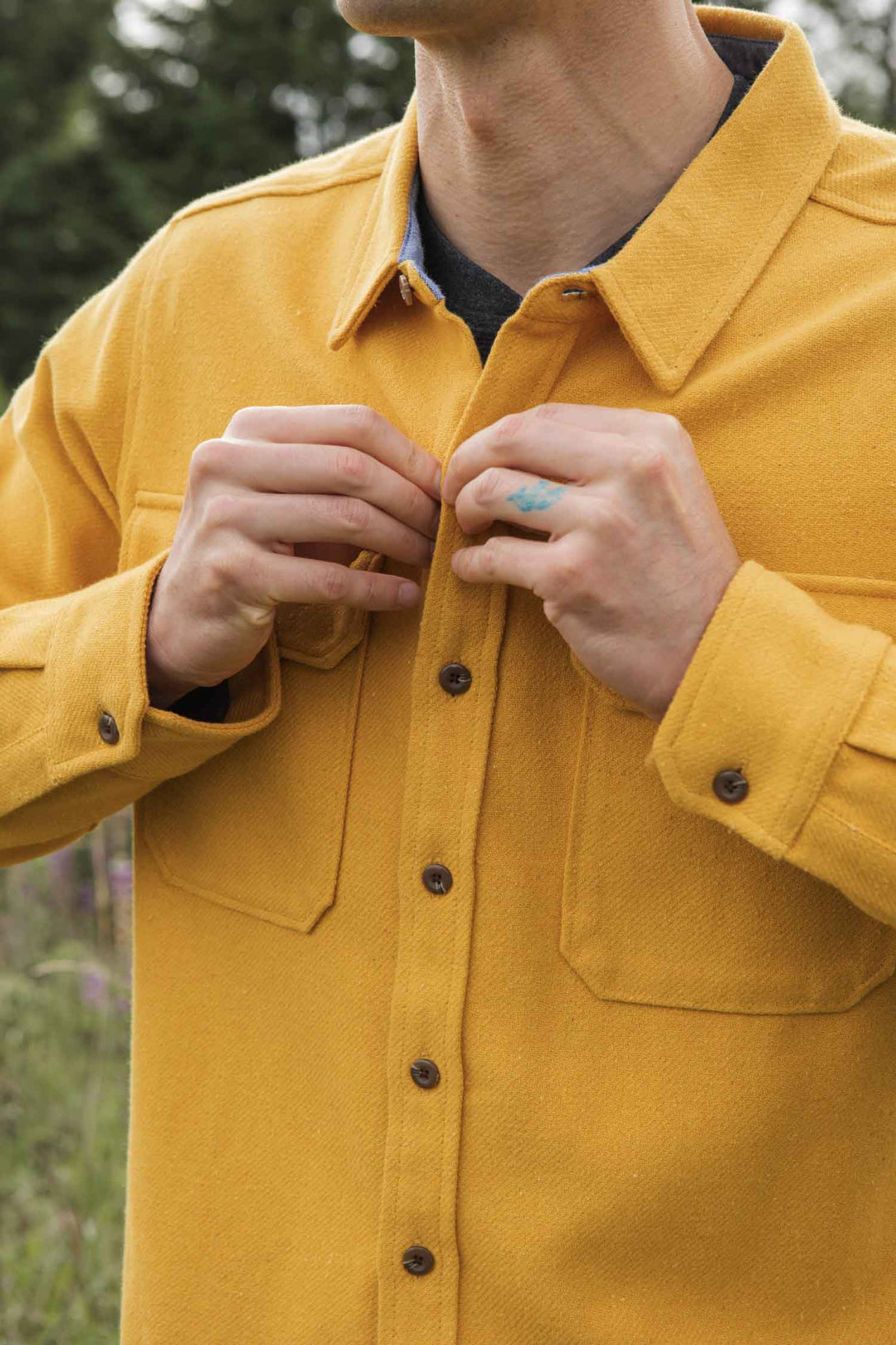 A person fastens the buttons of the Bridge & Burn Pointer Overshirt in Antique Gold, made from recycled fibers. Only their upper body is visible against a backdrop of blurred greenery.