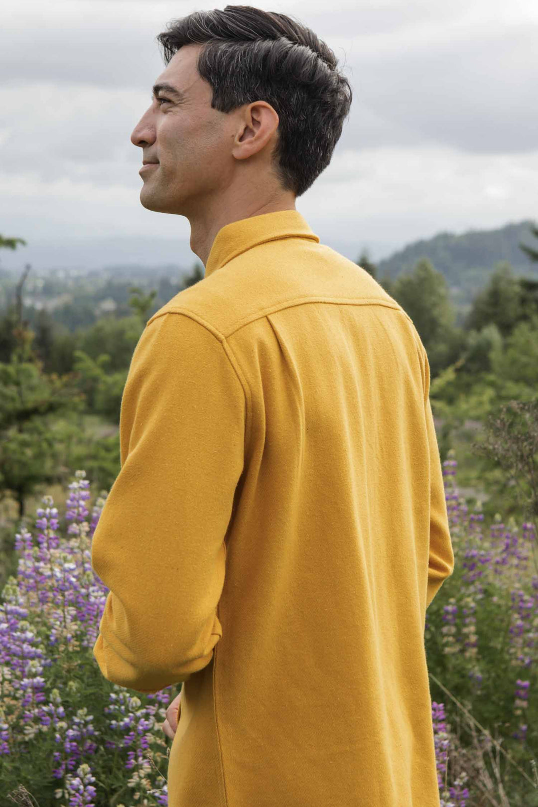 A man in the Bridge & Burn Pointer Overshirt in Antique Gold stands outdoors, mostly back to the camera, with purple wildflowers and green trees in the background beneath a cloudy sky.