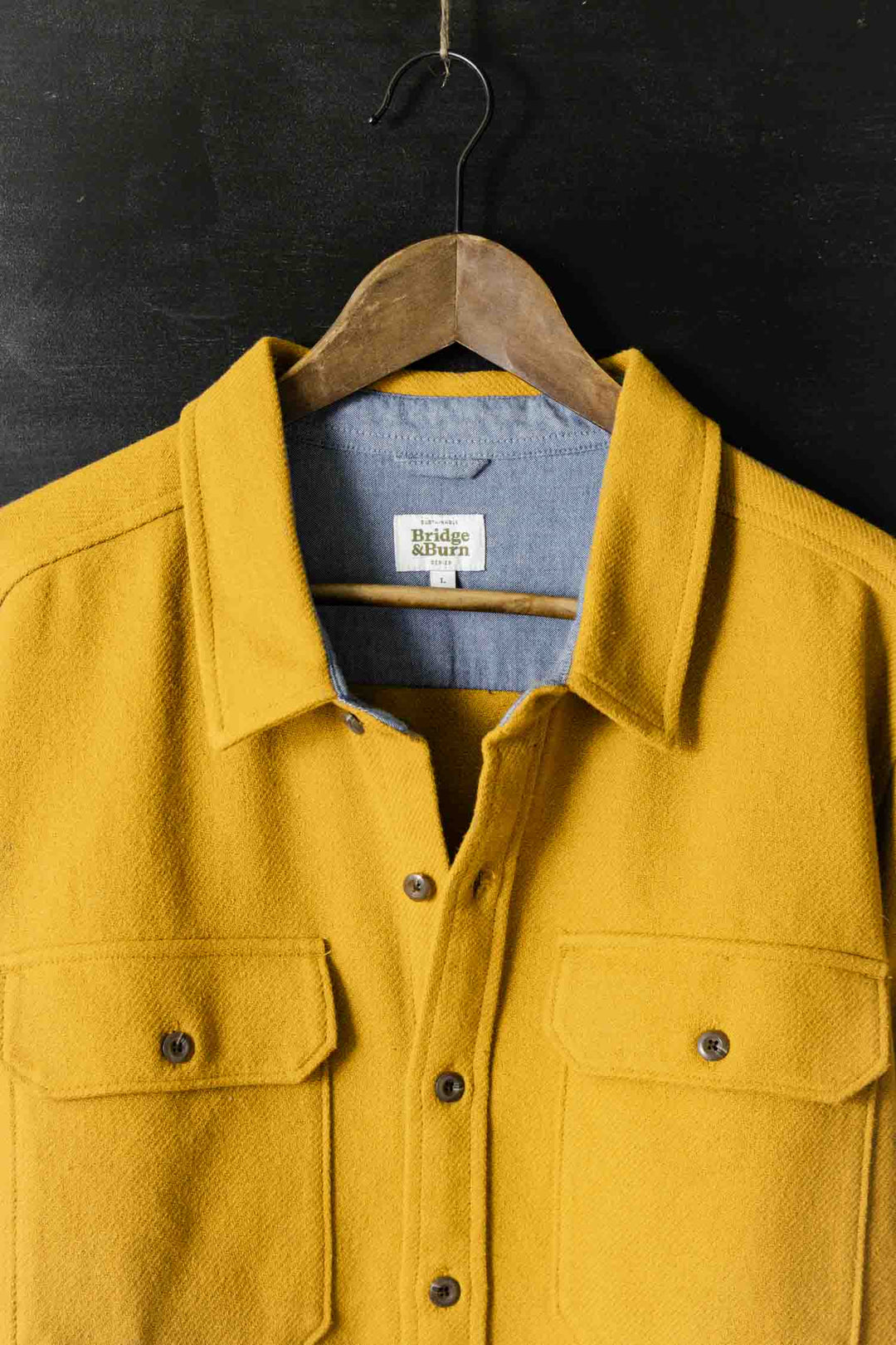 The Bridge & Burn Pointer Overshirt in Antique Gold hangs on a wooden hanger against a dark background, featuring front pockets, a light blue inner lining made from recycled fibers, and a visible label inside the collar.