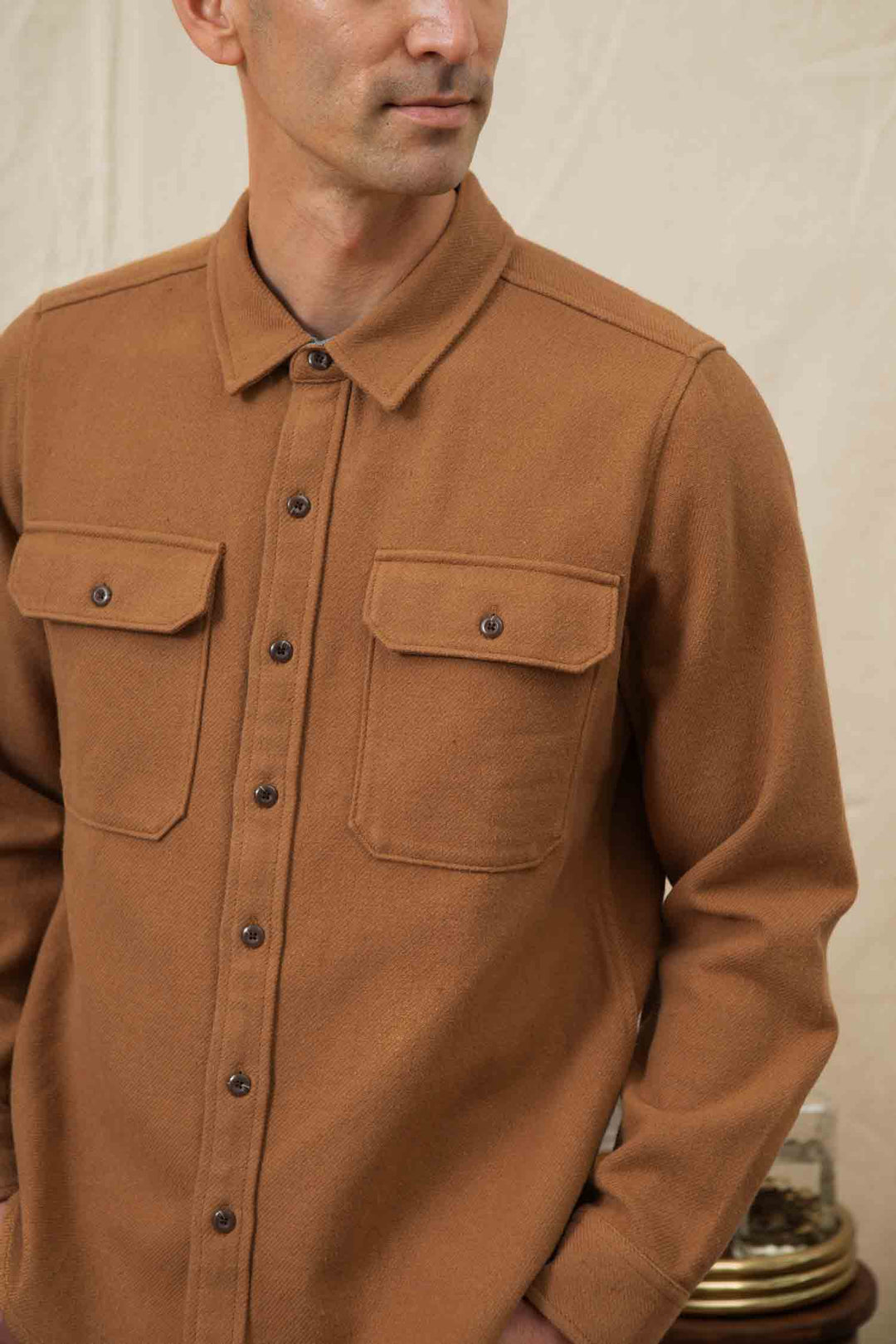 A man models the Bridge & Burn Pointer Overshirt in camel, featuring two chest pockets and a sustainable design. He stands against a neutral background, looking slightly to the side with only his upper body visible.