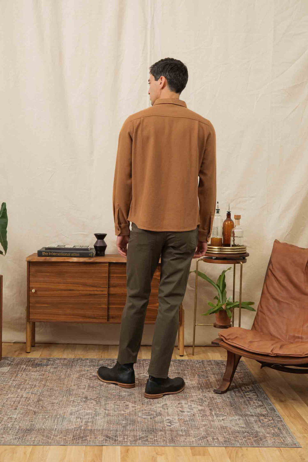 A man stands indoors with his back to the camera, wearing the Bridge & Burn Pointer Overshirt in Camel and olive green pants, in a room with mid-century modern furniture, plants, and neutral decor.