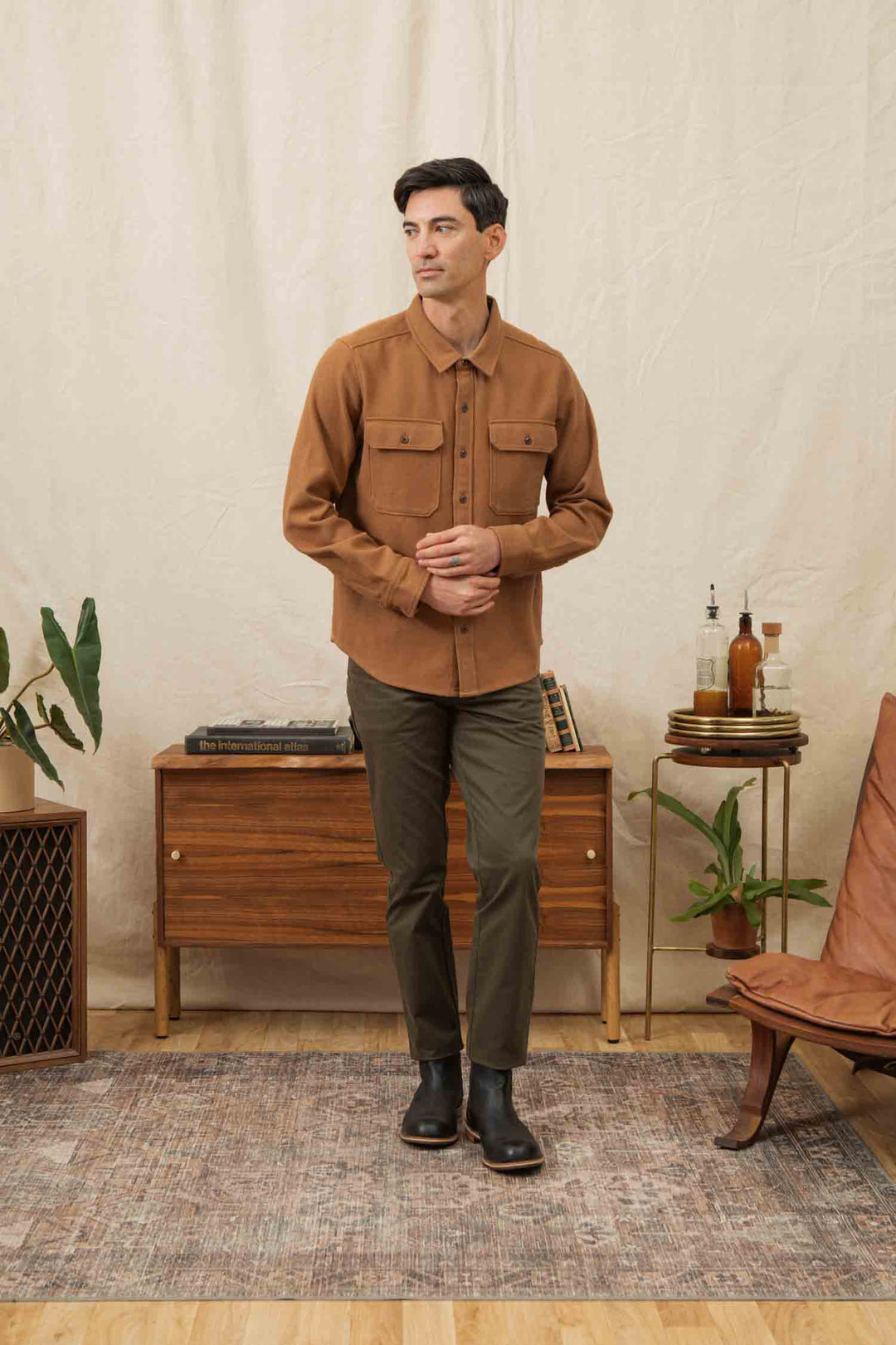 A man stands indoors on a patterned rug, wearing the Bridge & Burn Pointer Overshirt in Camel and olive pants. Behind him are mid-century wooden furniture, plants, books, bottles, and a cream-colored backdrop.
