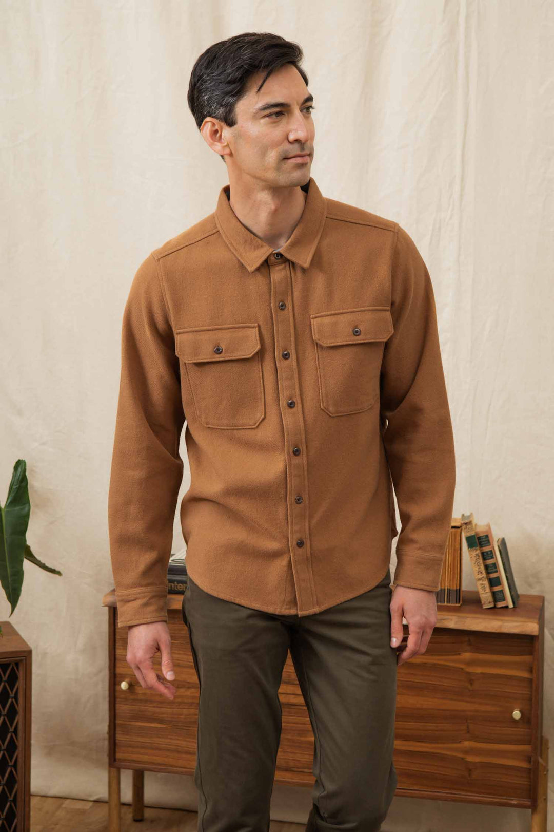 A man with short dark hair wears the Bridge & Burn Pointer Overshirt in camel—an eco-friendly brown button-up with two chest pockets—and olive green pants, standing indoors by a wooden cabinet and cream-colored curtain.
