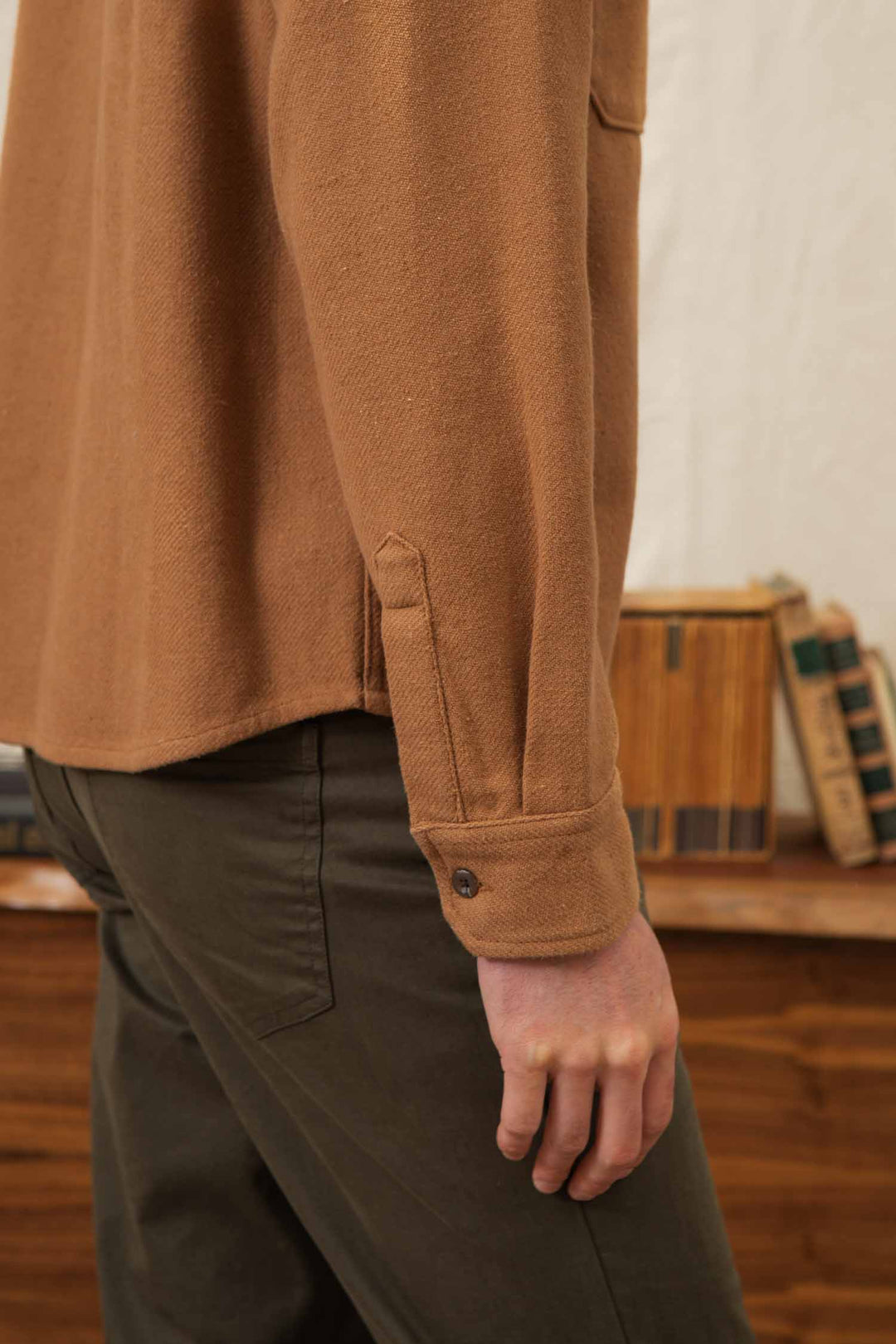 Someone wearing the Bridge & Burn Pointer Overshirt in Camel and olive green pants stands indoors near a wooden table with stacked vintage books, showing only their torso and arm.