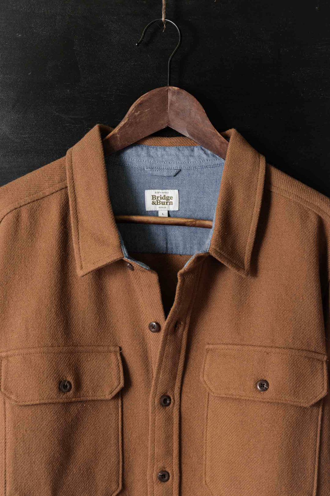 The Bridge & Burn Pointer Overshirt in Camel, made from recycled fibers, hangs on a wooden hanger against a dark background. Featuring two front pockets, collar, and visible neck tag, it’s a sustainable and stylish layering piece.