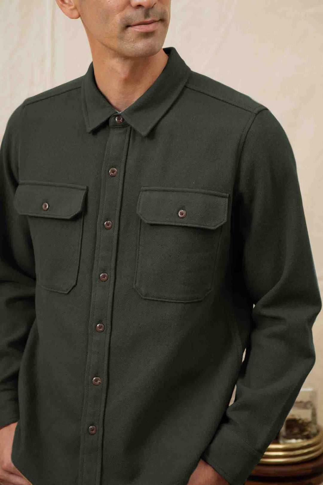 A person wears the Bridge & Burn Pointer Overshirt in Dark Sage, made from recycled fibers with two chest pockets, standing against a neutral background. Only the upper body and part of the face are visible.