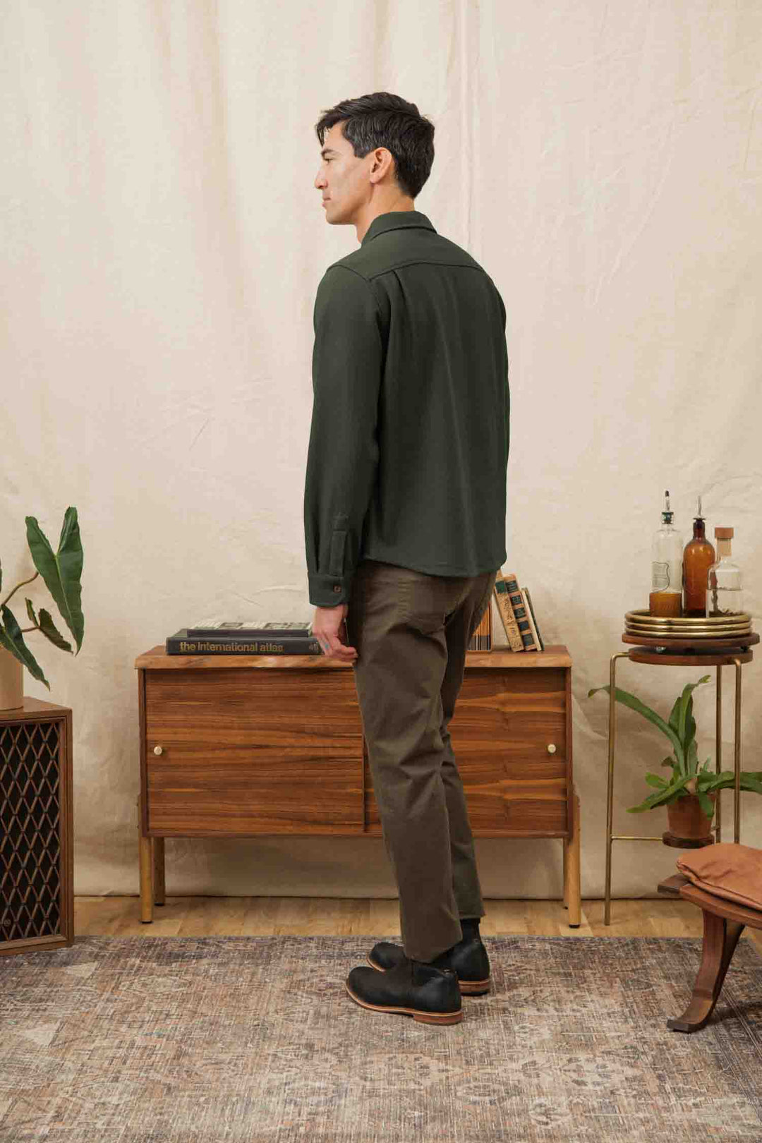 A man stands indoors with his back to the camera, wearing the Bridge & Burn Pointer Overshirt in Dark Sage and brown pants. He is in a warm, cozy room with a wooden sideboard, books, plants, and bottles—reflecting sustainable style.