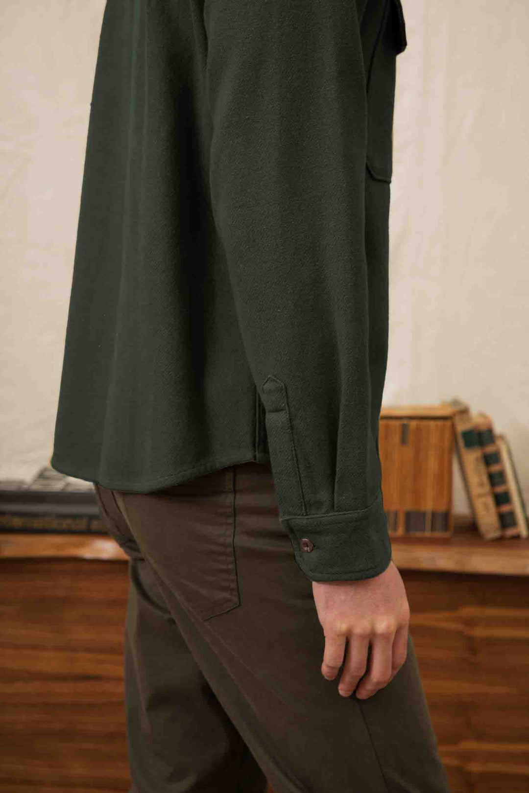 A person wears the Bridge & Burn Pointer Overshirt in Dark Sage with brown pants indoors; their side and arm are visible near a wooden table stacked with books, evoking a cozy, sustainability-focused atmosphere.