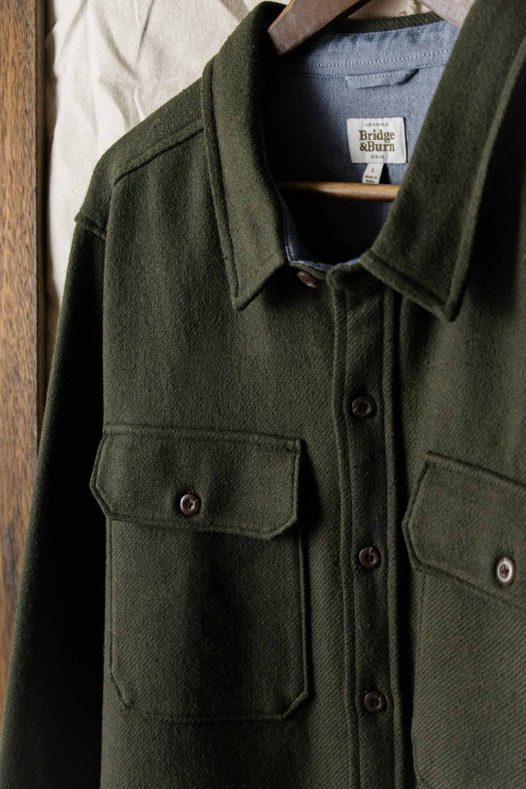 The Bridge & Burn Pointer Overshirt in Dark Sage, made from thick, textured twill, features two chest pockets and hangs on a hanger against a wood and fabric background.