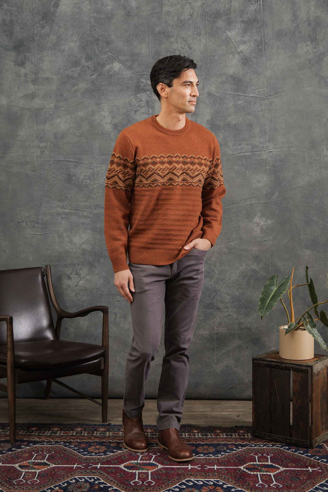 A man stands indoors on a patterned rug wearing the Bridge & Burn Rockwood Fairisle Sweater in Walnut Grove, gray pants, and brown shoes. He has one hand in his pocket and looks to the side, with a chair and potted plant nearby.