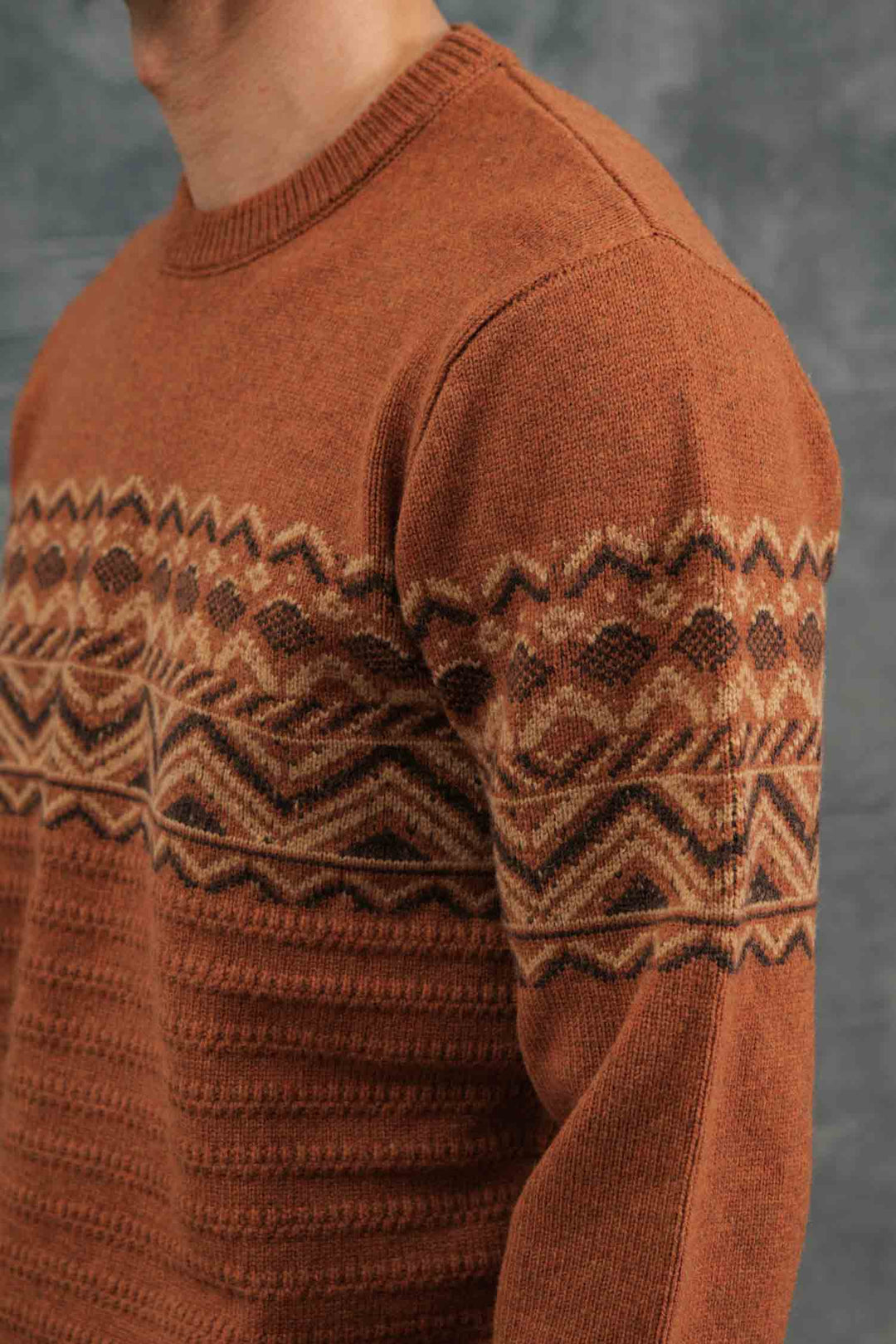 A person wears the Bridge & Burn Rockwood Fairisle Sweater in Walnut Grove, featuring RWS certified merino wool and a cream and brown geometric zigzag pattern, standing against a gray background.