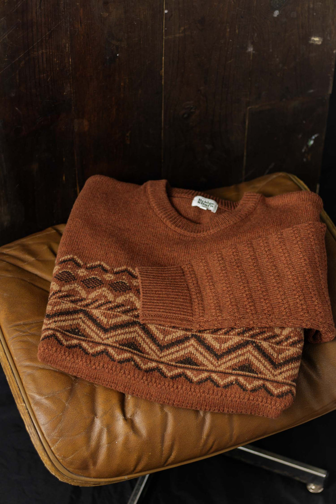 The Rockwood Fairisle Sweater in Walnut Grove by Bridge & Burn, featuring a geometric zigzag pattern in earthy tones, is folded on a worn tan leather chair set against a dark wooden backdrop.