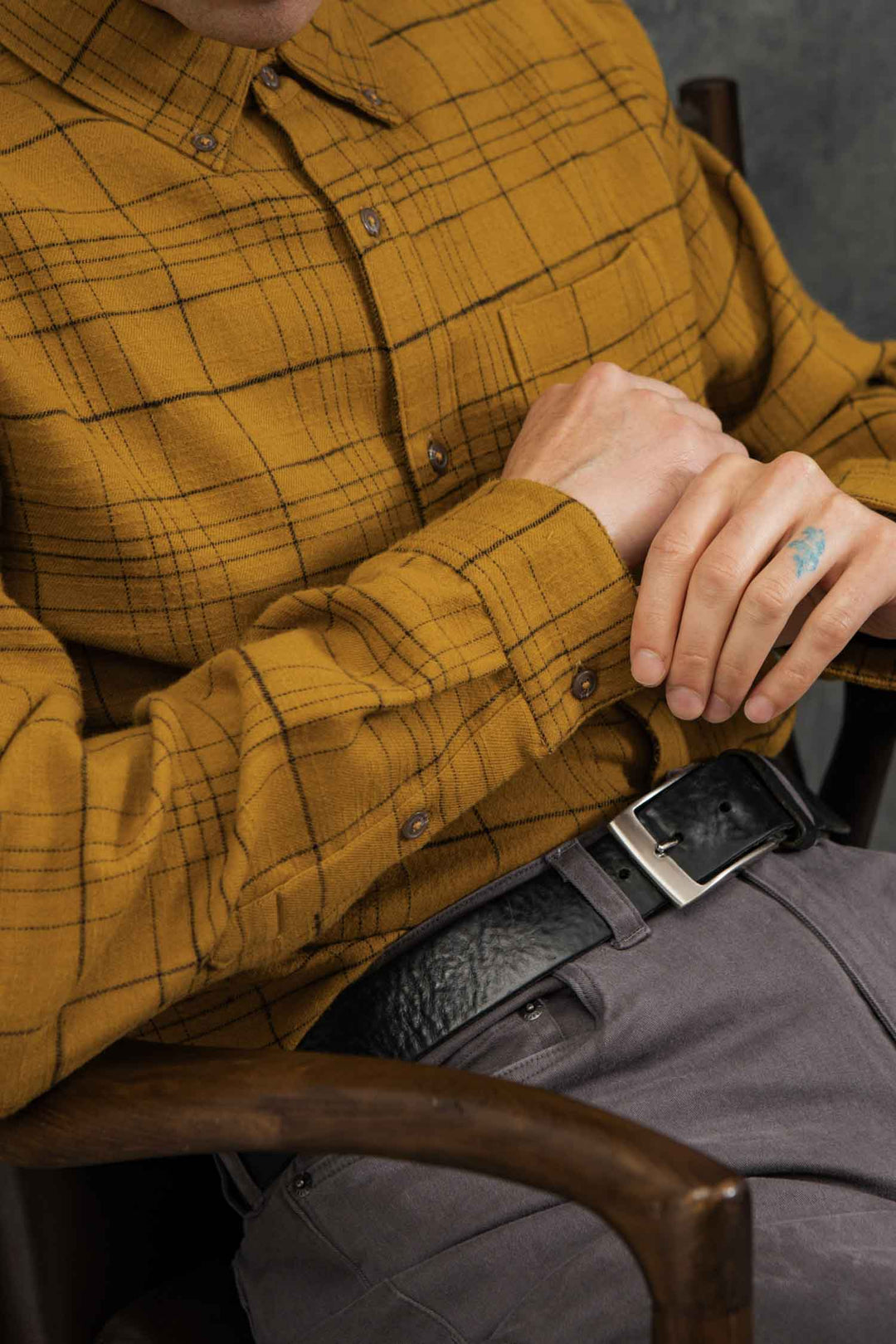 A person wearing the Bridge & Burn Sutton Slim Shirt in Golden Horizon Plaid with gray pants sits hands clasped, showing a blue hand tattoo and a black belt with silver buckle. The face is partially out of frame.