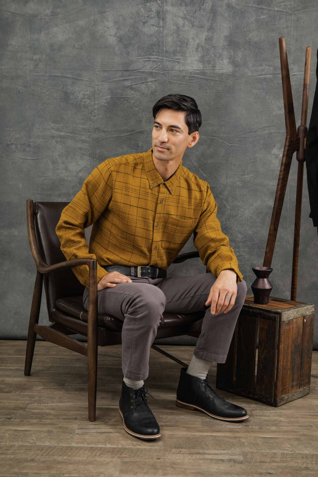 A man with dark hair wears the Bridge & Burn Sutton Slim Shirt in Golden Horizon Plaid and gray pants as he sits on a leather chair, looking to the side. A coat rack and wooden box complement the minimalist decor nearby.