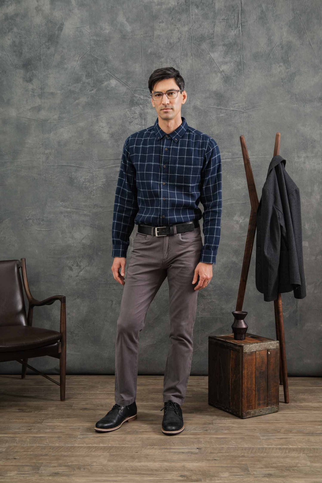 A man wearing glasses, gray pants, and black shoes stands indoors on a wooden floor. He is wearing the Bridge & Burn Sutton Slim Shirt in Moonrise Windowpane. Behind him are a chair, a coat rack with a jacket, a box, and a gray backdrop.