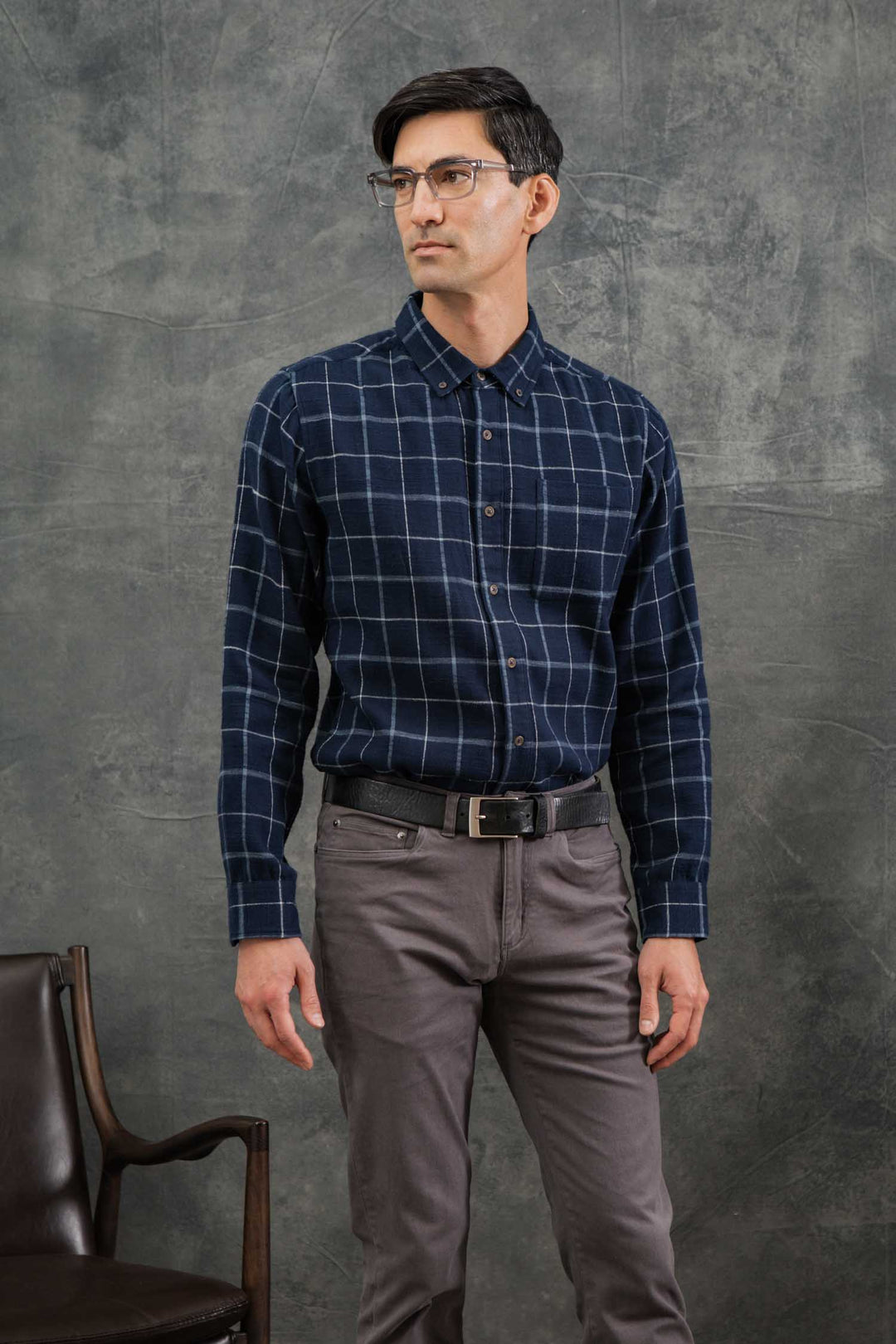 A man wearing glasses and the Bridge & Burn Sutton Slim Shirt in Moonrise Windowpane stands indoors near a brown chair, looking to the side against a gray textured background.