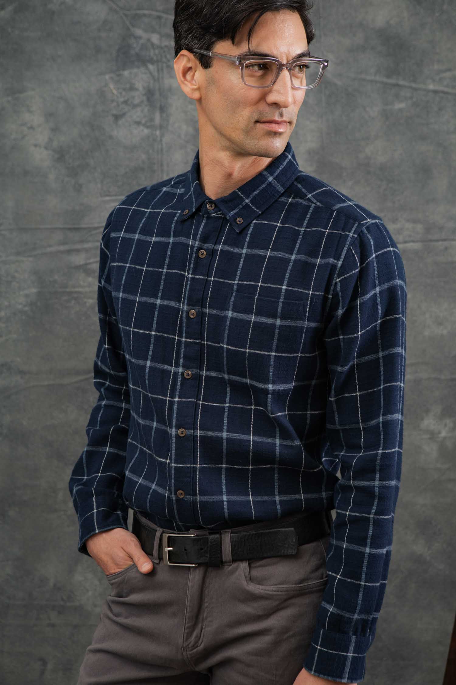 A man models the Bridge & Burn Sutton Slim Shirt in Moonrise Windowpane with gray pants against a textured gray background, one hand in his pocket and looking confidently to the side.