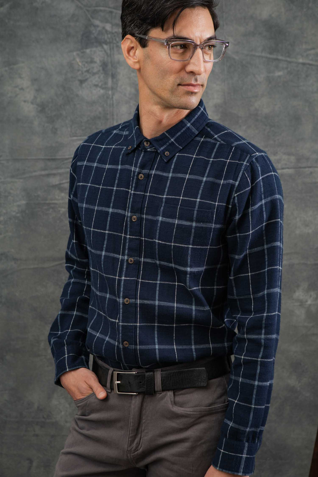 A man models the Bridge & Burn Sutton Slim Shirt in Moonrise Windowpane with gray pants against a textured gray background, one hand in his pocket and looking confidently to the side.