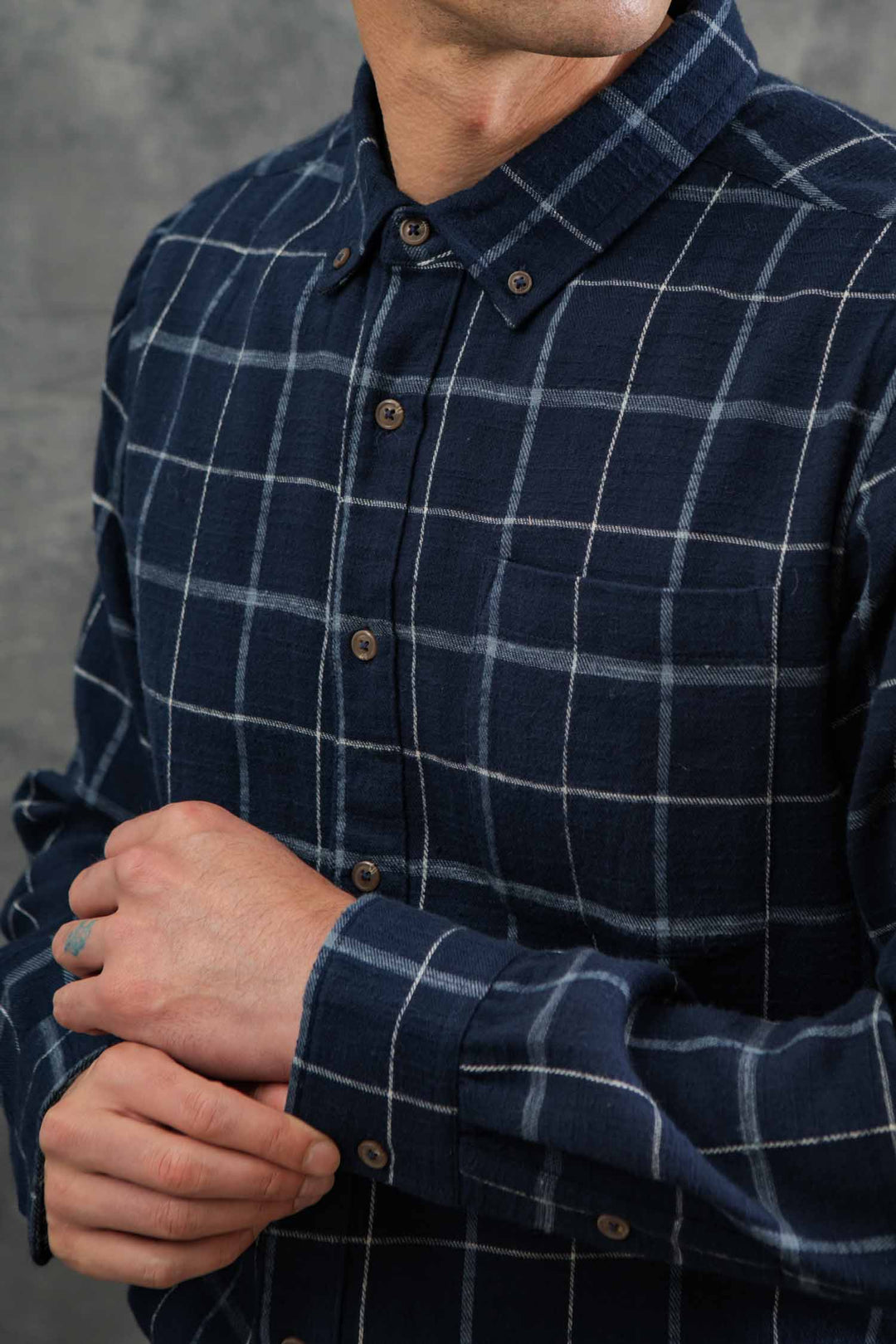 Wearing the Bridge & Burn Sutton Slim Shirt in Moonrise Windowpane, a person adjusts their left cuff with their right hand against a gray background, highlighting the shirt's dark blue plaid and minimalist design. Face is partially out of frame.