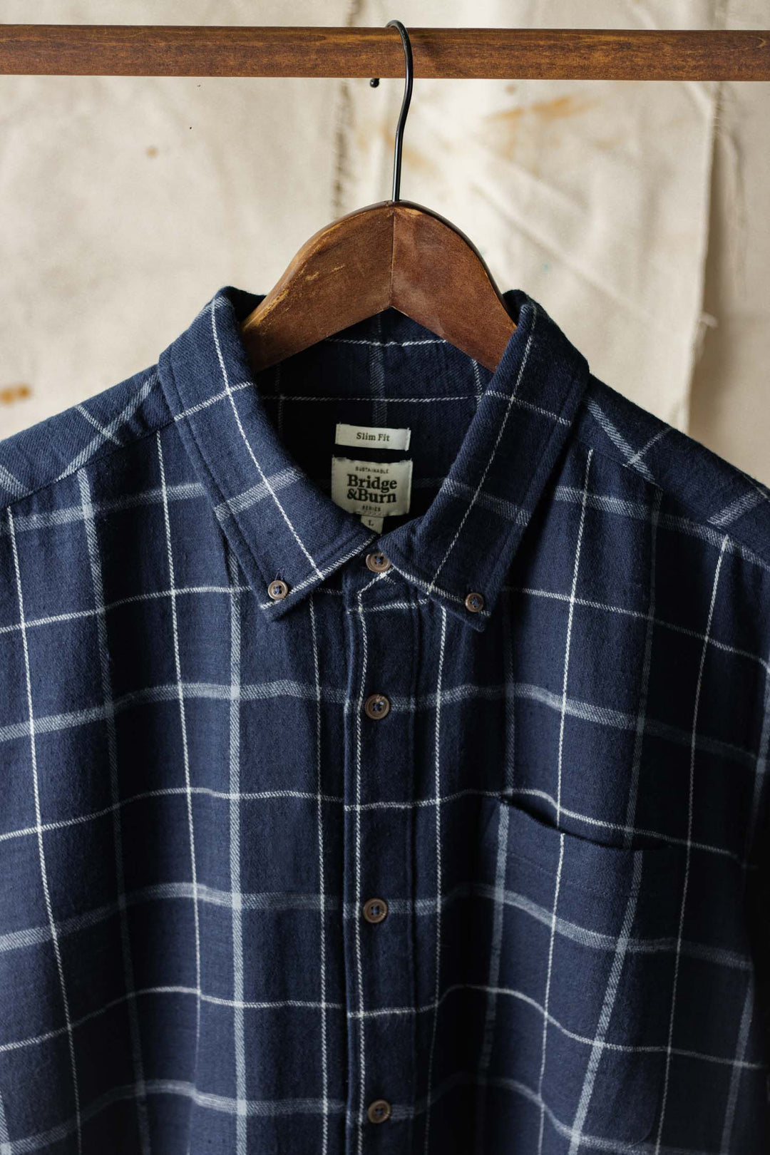 The Bridge & Burn Sutton Slim Shirt in Moonrise Windowpane, a navy blue slim fit flannel with white check pattern, pointed collar, and chest pocket, is displayed on a wooden hanger against a textured neutral background.