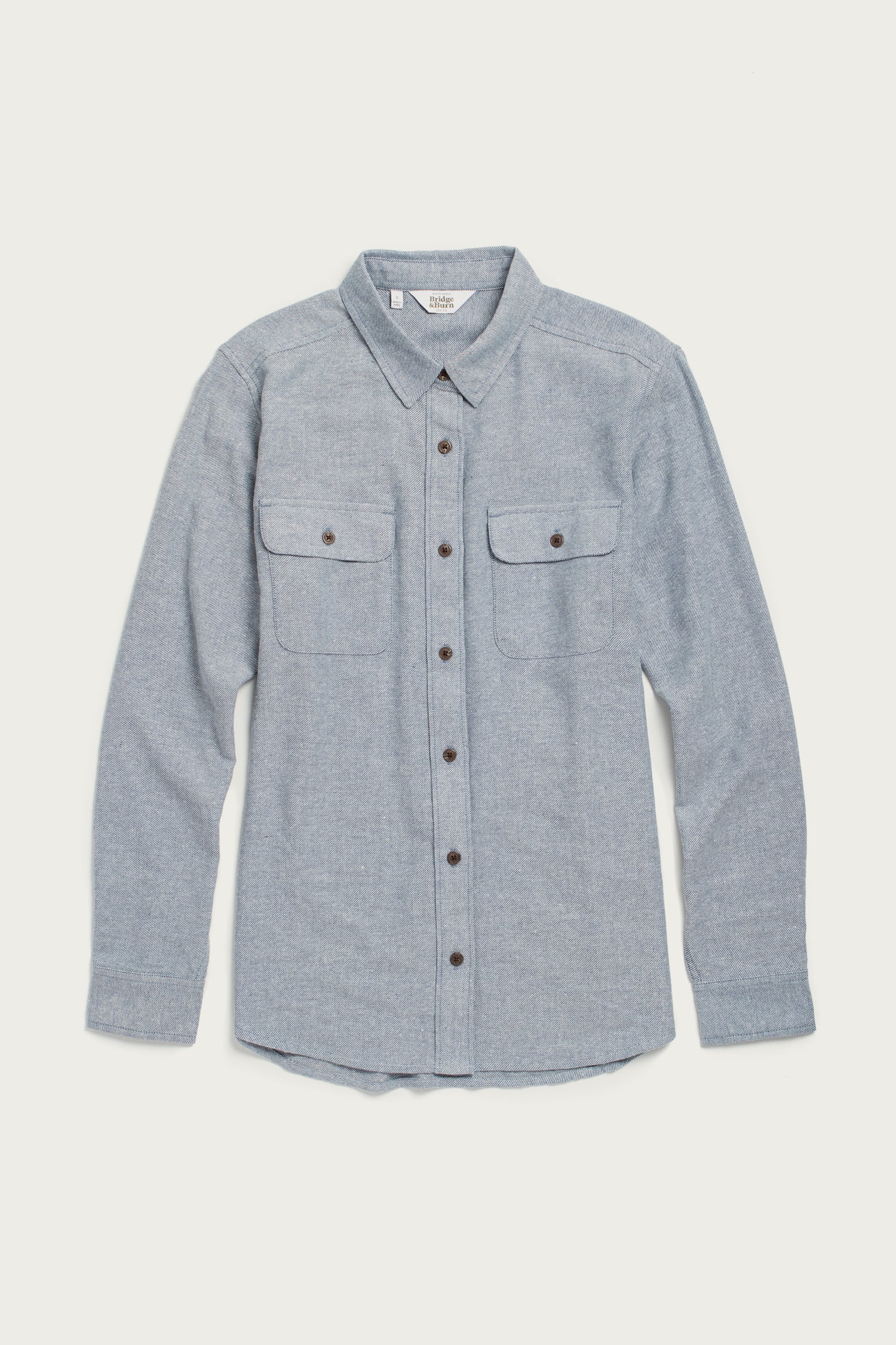 The Bridge & Burn Lenon Shirt in Denim Twill is a light blue, long-sleeve, relaxed-fit collared shirt with two chest pockets, made from recycled cotton and shown laid flat on a white background.