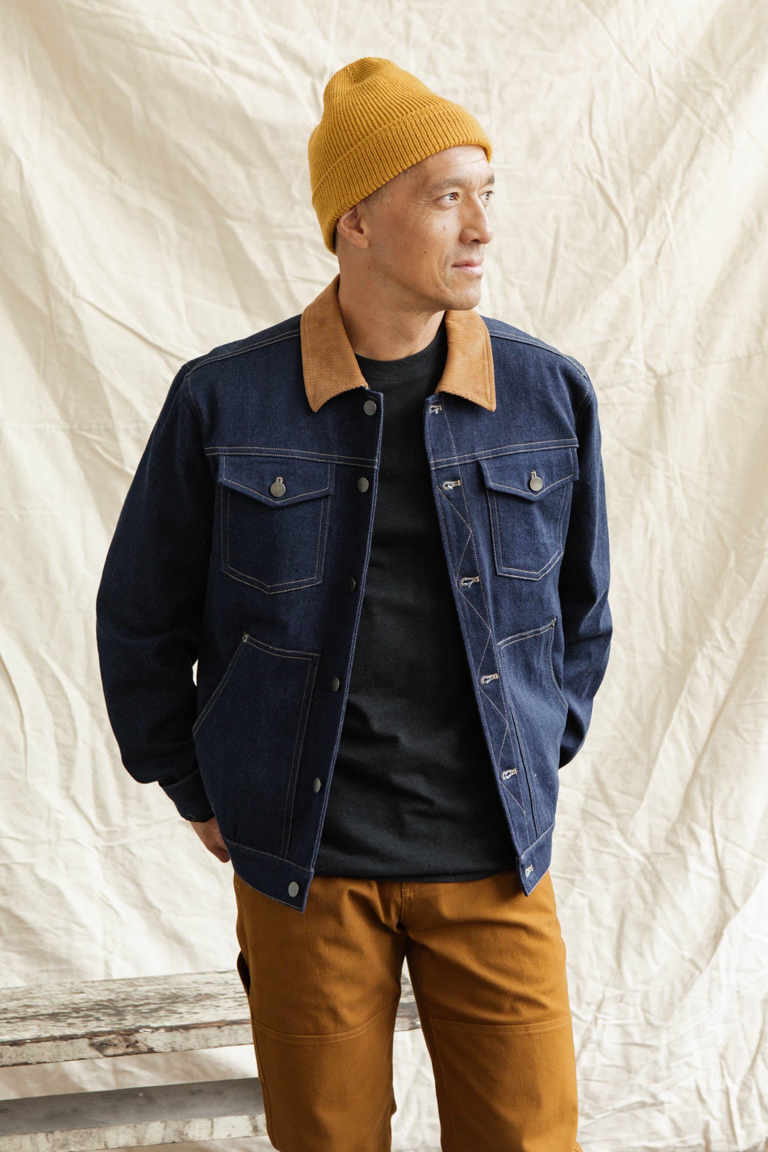 A person in the Bridge & Burn Burl Trucker Jacket in Dark Denim with a tan corduroy collar, a mustard-yellow beanie, black shirt, and mustard-brown pants stands before a beige fabric backdrop, looking to the side and smiling.