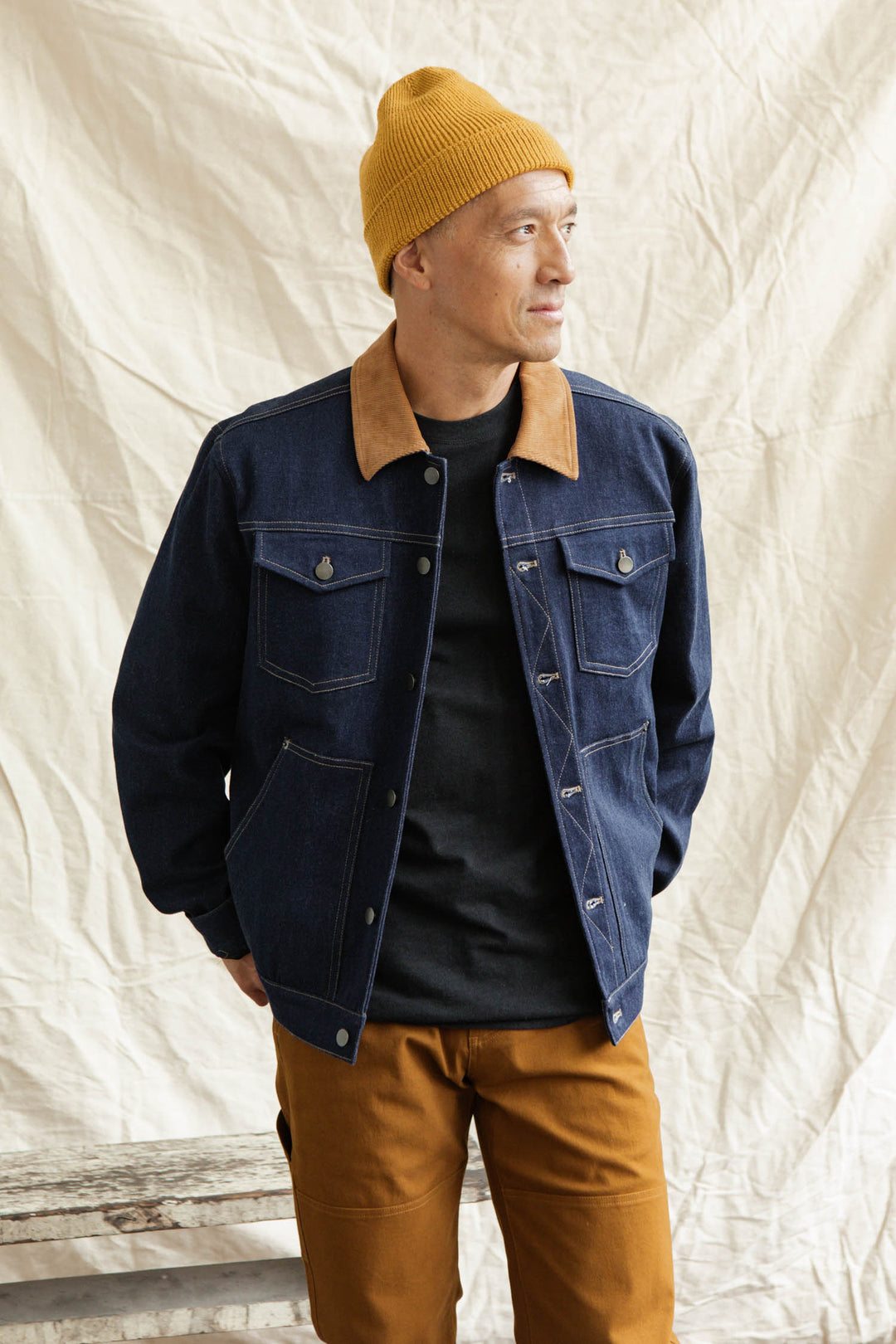 A person in the Bridge & Burn Burl Trucker Jacket in Dark Denim with a tan corduroy collar, a mustard-yellow beanie, black shirt, and mustard-brown pants stands before a beige fabric backdrop, looking to the side and smiling.
