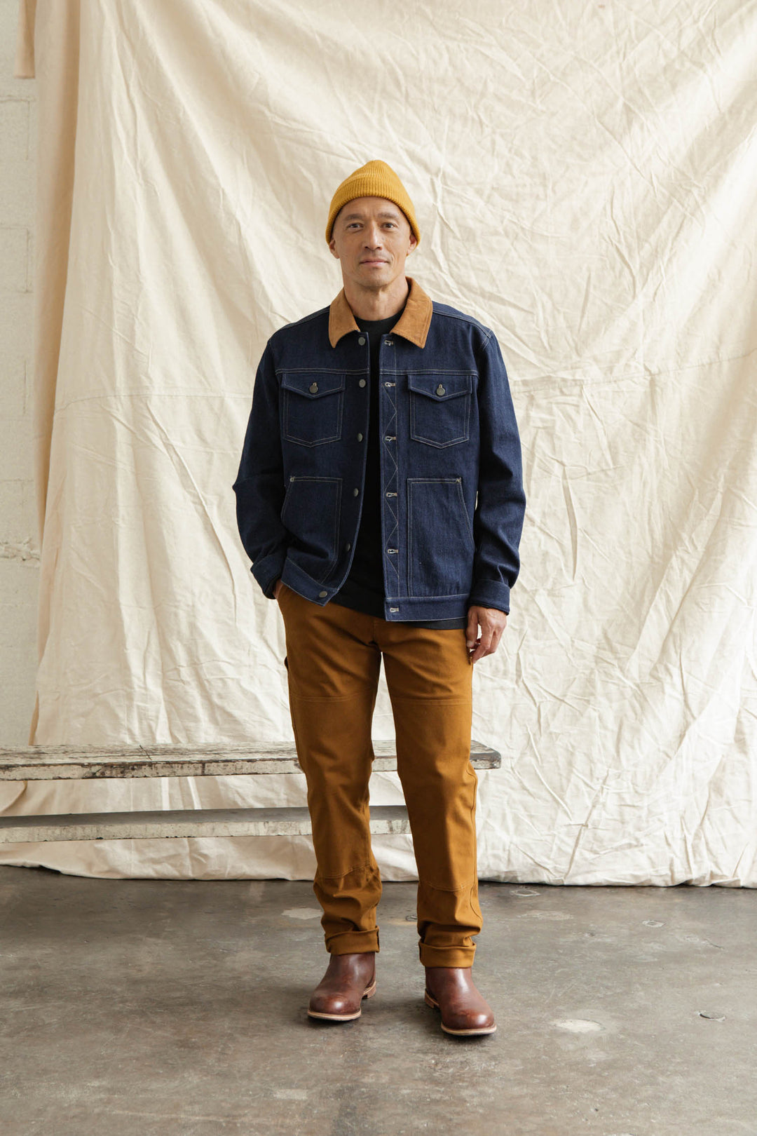 Indoors before cream drapes, a person wears the Bridge & Burn Burl Trucker Jacket in Dark Denim with tan corduroy trim, yellow beanie, black shirt, brown pants, and brown boots, hands tucked in their jacket pockets.