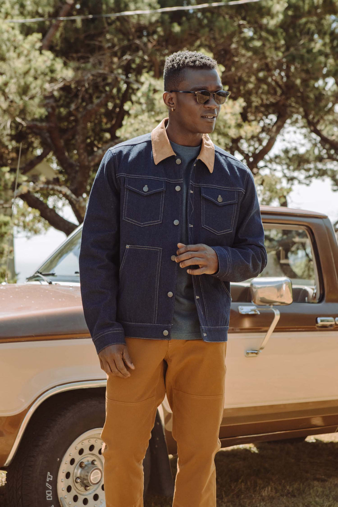 A man wearing sunglasses and the Bridge & Burn Burl Trucker Jacket in Dark Denim stands outdoors in front of a vintage brown pickup truck with trees in the background.
