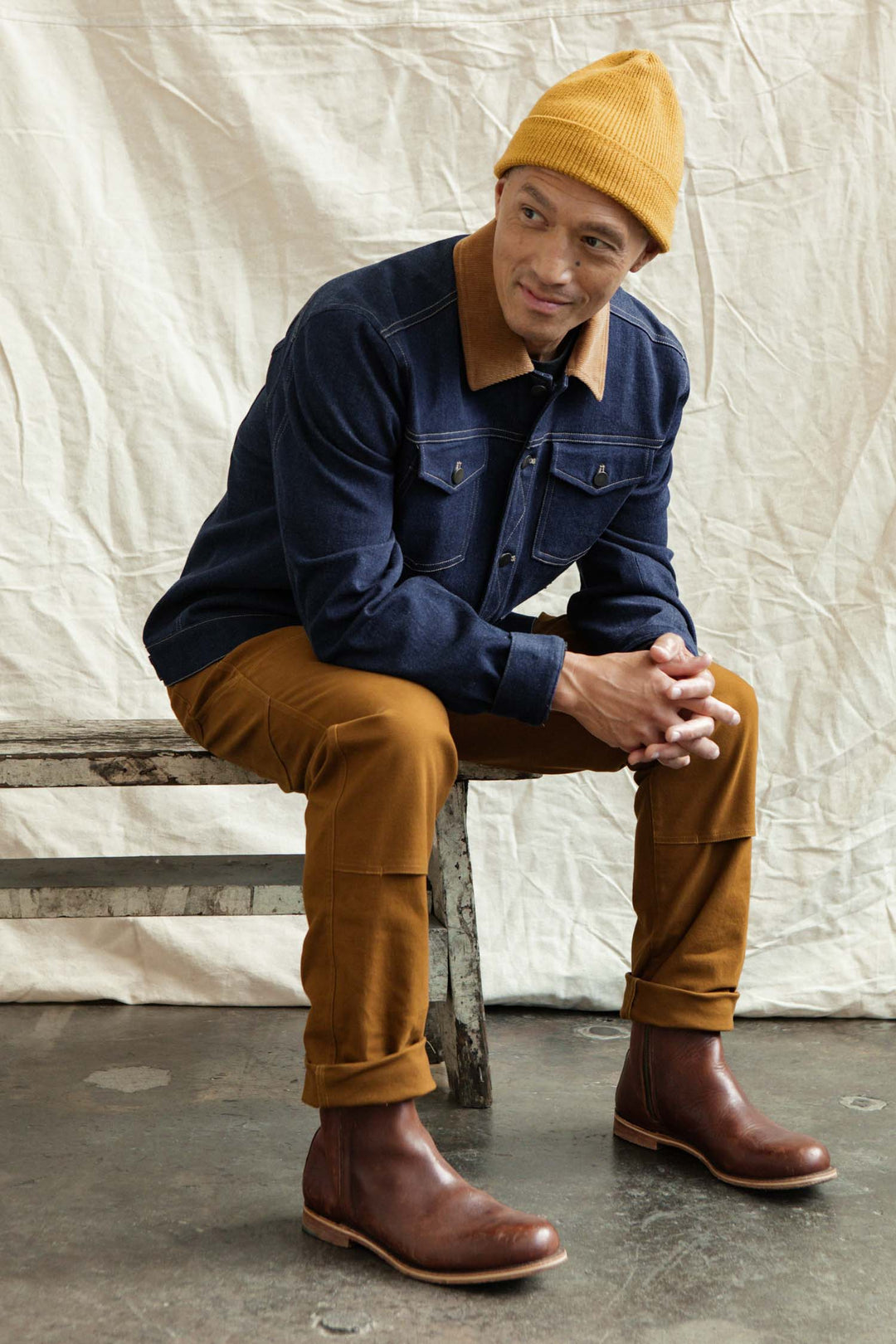 A man in a mustard yellow beanie, Bridge & Burn Burl Trucker Jacket in dark denim, mustard pants, and brown boots sits on a rustic bench against a cream fabric backdrop, looking slightly to his left with hands clasped.