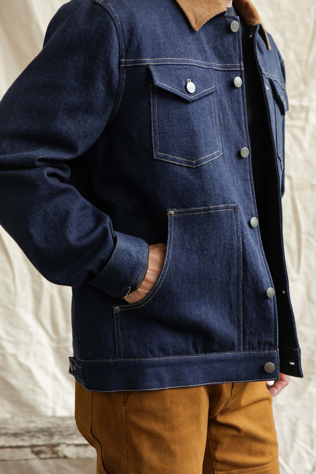 A person models the Bridge & Burn Burl Trucker Jacket in Dark Denim, featuring a brown corduroy collar, paired with brown pants. They stand sideways with one hand in their pocket against a neutral, blurred background.