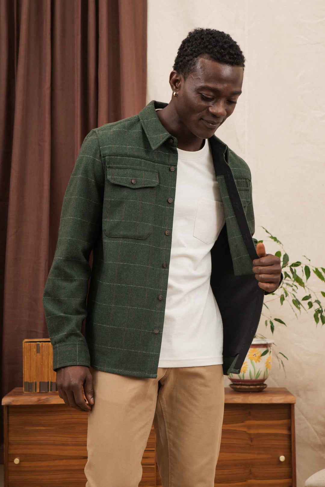 A man wearing the Bridge & Burn Fielding Shirt Jacket in Cypress Windowpane over a white tee and beige pants stands indoors, looking down and slightly opening his jacket. A plant and wooden furniture are visible behind him.