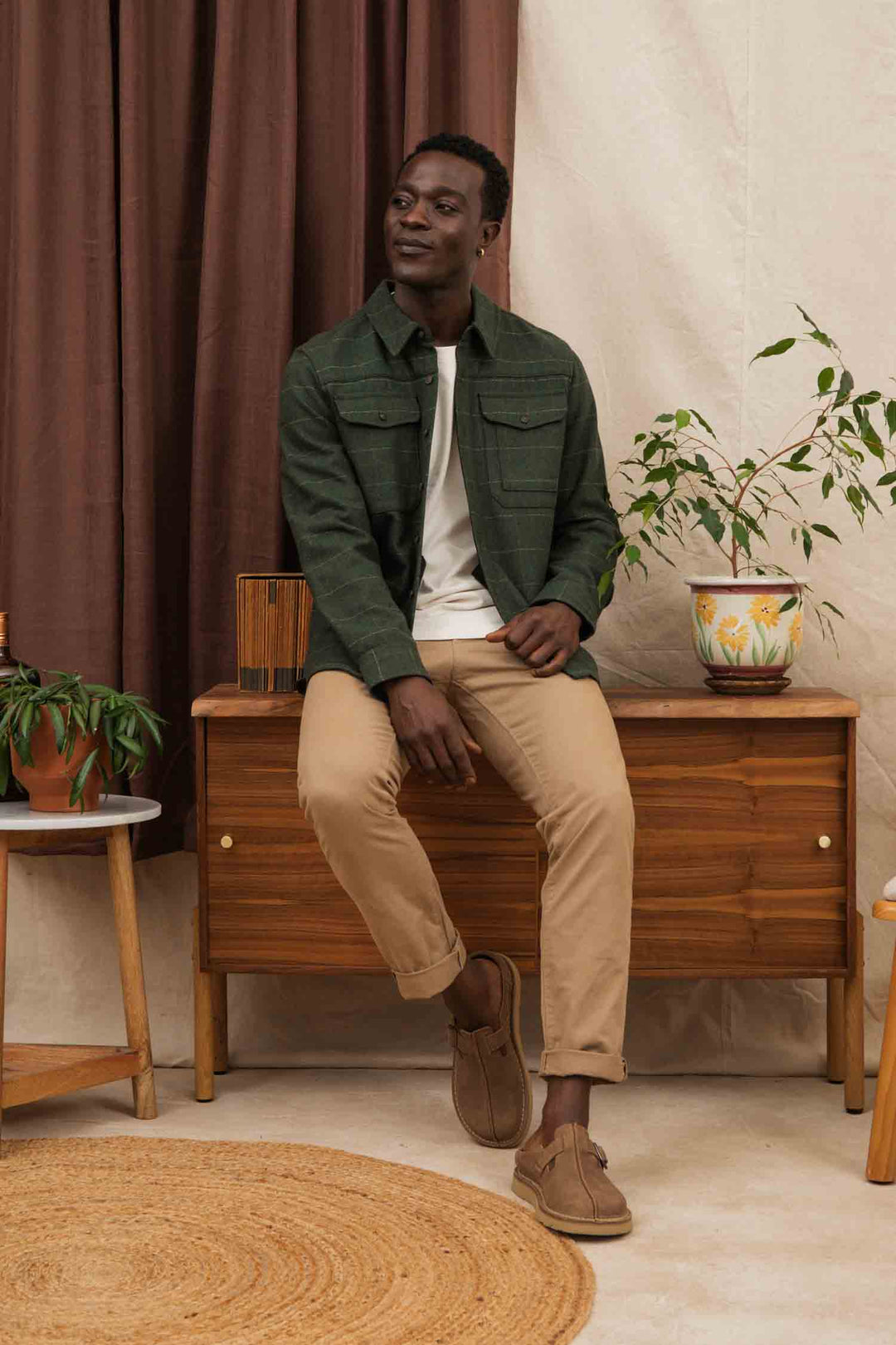 A man wearing the Bridge & Burn Fielding Shirt Jacket in Cypress Windowpane, a white shirt, and beige pants sits on a wooden cabinet beside potted plants in a cozy room with brown and cream curtains.