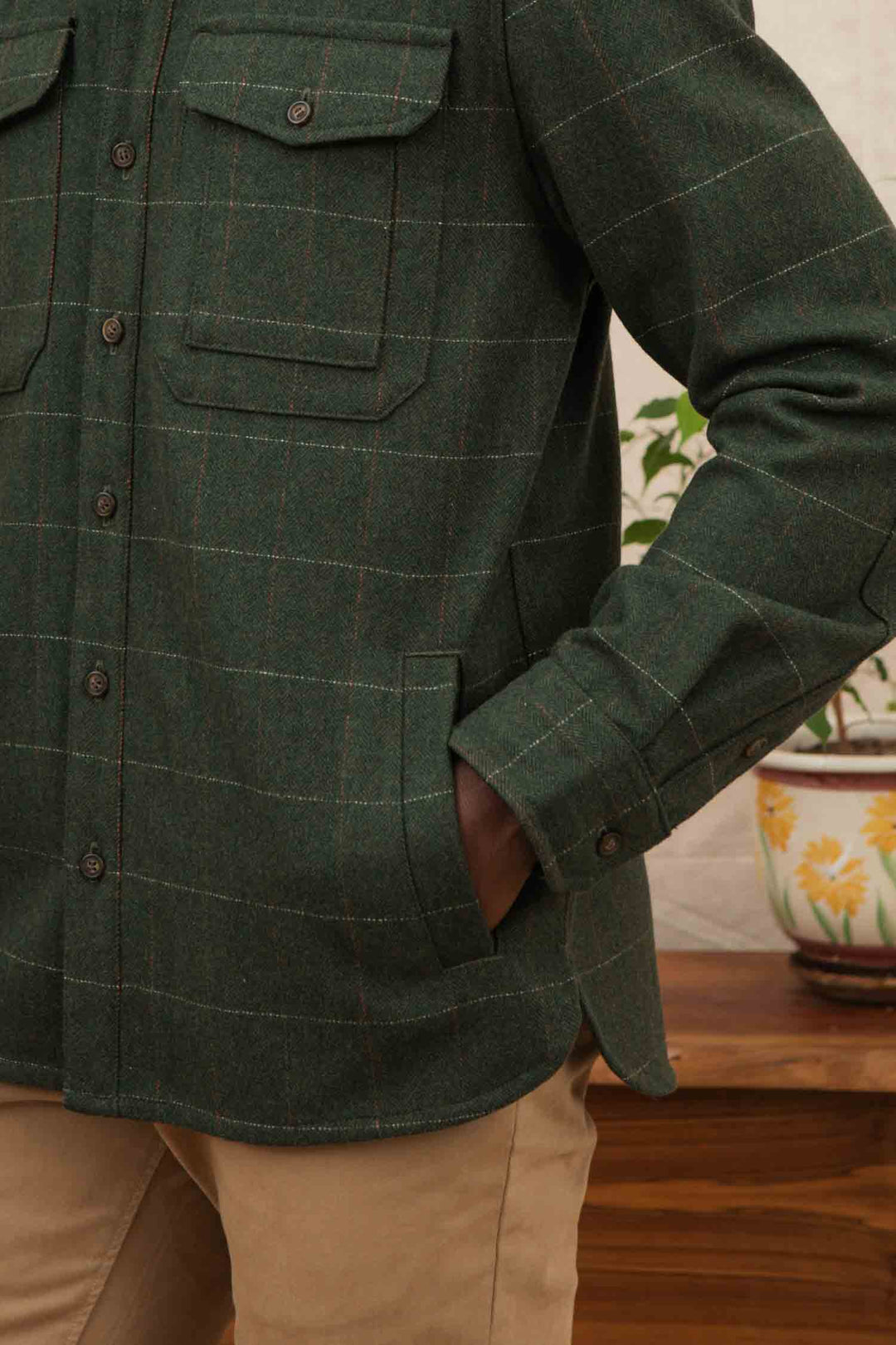 A person wears the Bridge & Burn Fielding Shirt Jacket in Cypress Windowpane with tan pants, one hand in their pocket. In the background, a plant in a floral pot is set on a wooden surface.