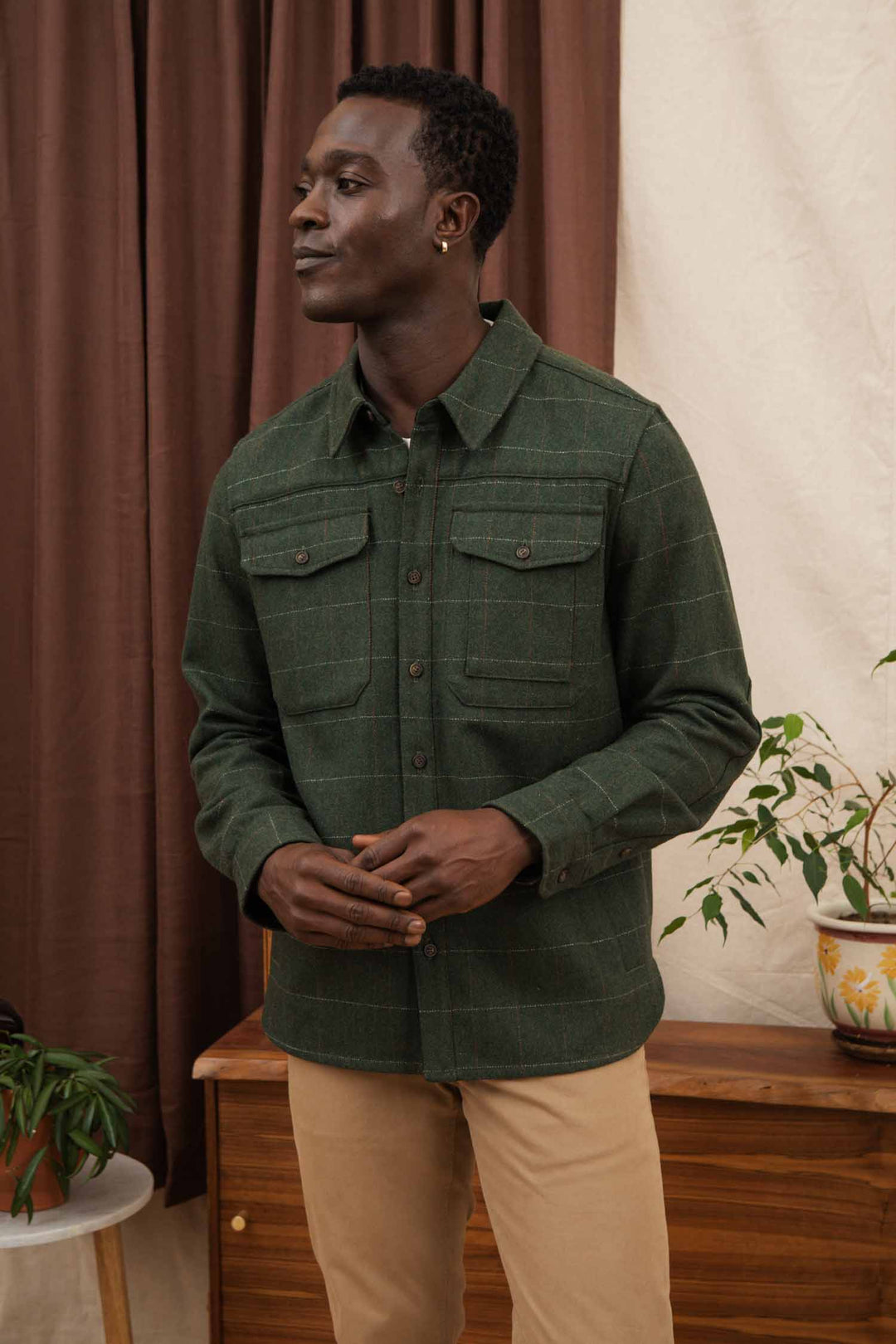 A man stands indoors in front of brown curtains and a wooden cabinet with potted plants, wearing the Bridge & Burn Fielding Shirt Jacket in Cypress Windowpane and beige pants.