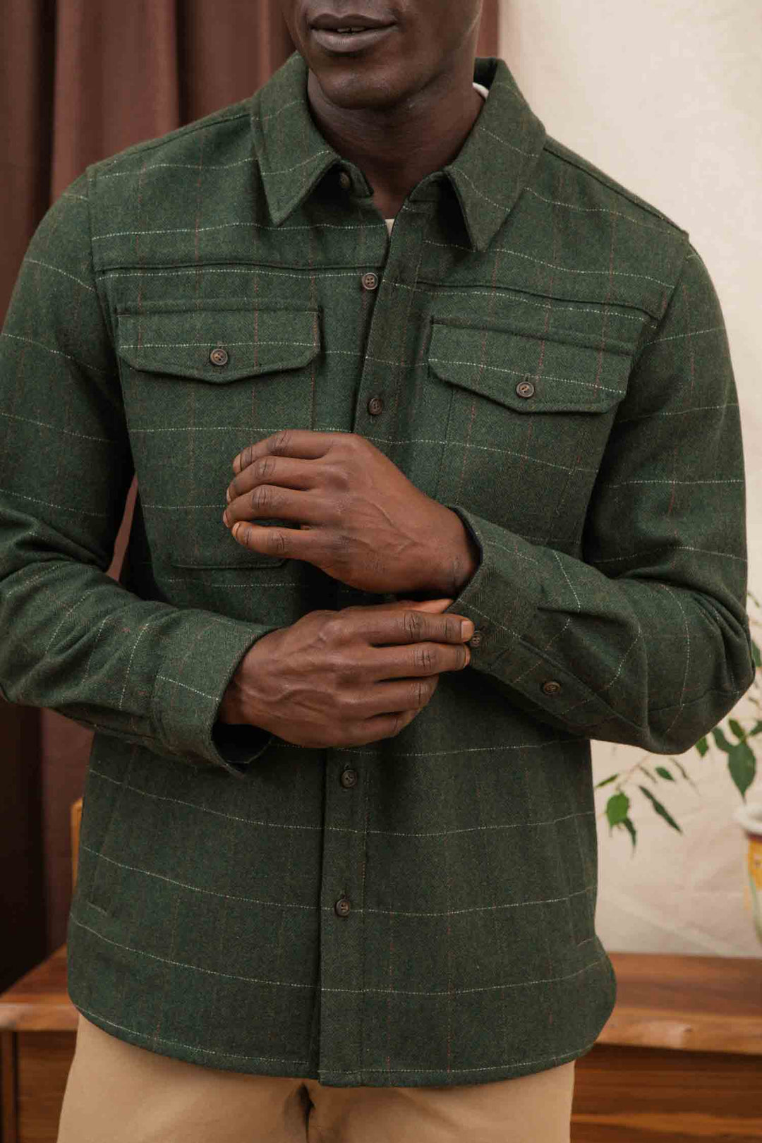 A man indoors wears the Bridge & Burn Fielding Shirt Jacket in Cypress Windowpane, featuring green plaid wool and two chest pockets, paired with beige pants. He adjusts his sleeve; only his torso and lower face are visible.