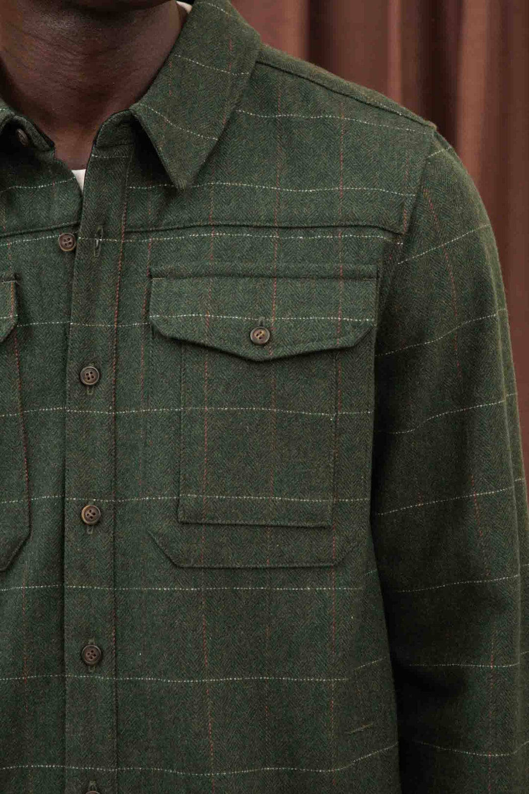 A close-up of a person wearing the Bridge & Burn Fielding Shirt Jacket in Cypress Windowpane—a green flannel with yellow and red grid lines and two chest pockets. Brown curtains are visible in the background.