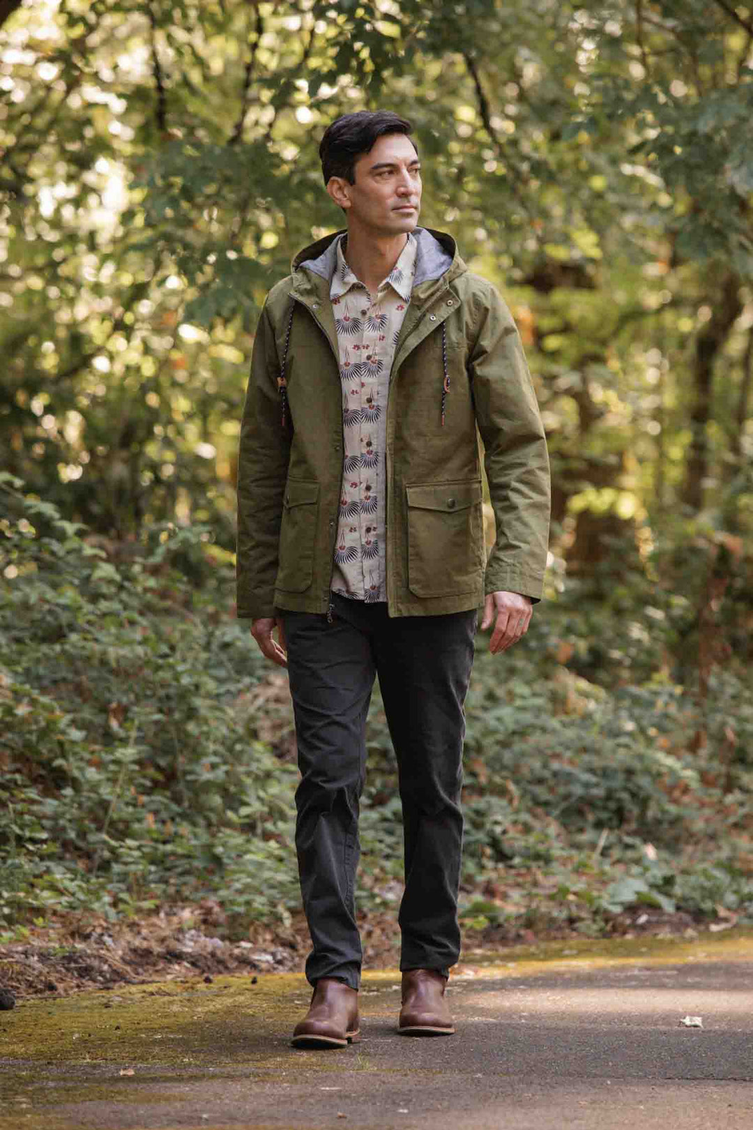 A man walks through a forest in the Bridge & Burn Marshall Jacket in Olive Waxed Canvas—water-resistant and hooded—paired with a patterned shirt, dark pants, and brown boots, surrounded by greenery under soft daylight.