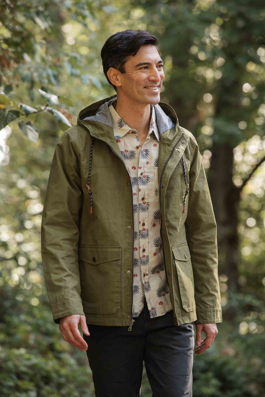 A man wearing the Bridge & Burn Marshall Jacket in Olive Waxed Canvas over a patterned shirt walks smiling through a lush green forest.