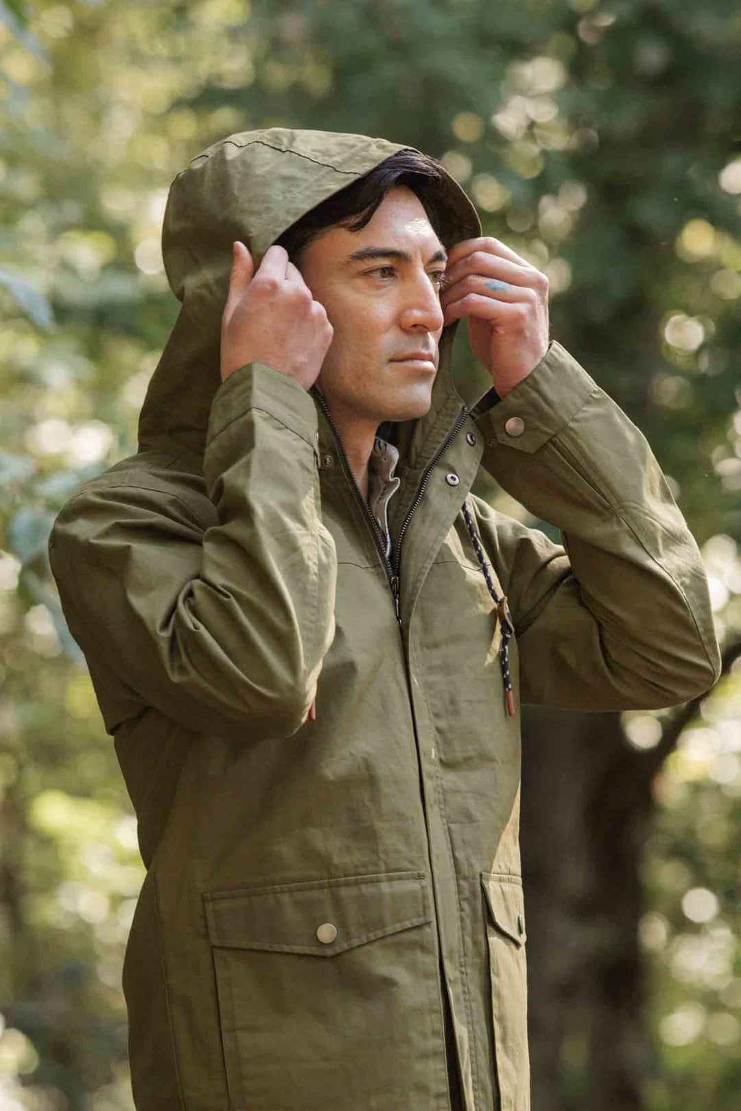 Wearing the Bridge & Burn Marshall Jacket in Olive Waxed Canvas, a person adjusts the hood outdoors amid blurred greenery and trees, highlighting the jacket’s water-resistant design and practical style for forest or park settings.