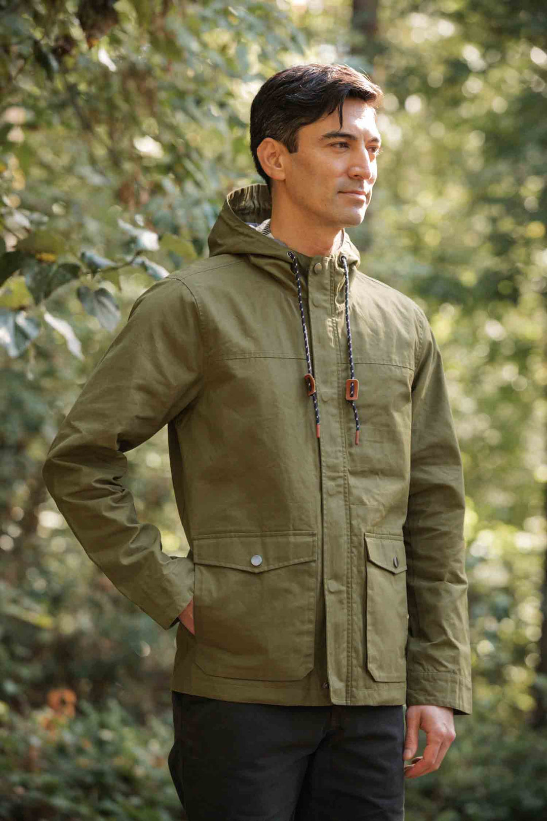 A man stands in a sunlit forest wearing the Bridge & Burn Marshall Jacket in olive waxed canvas, featuring a hood and front pockets, looking to the side with a relaxed expression.