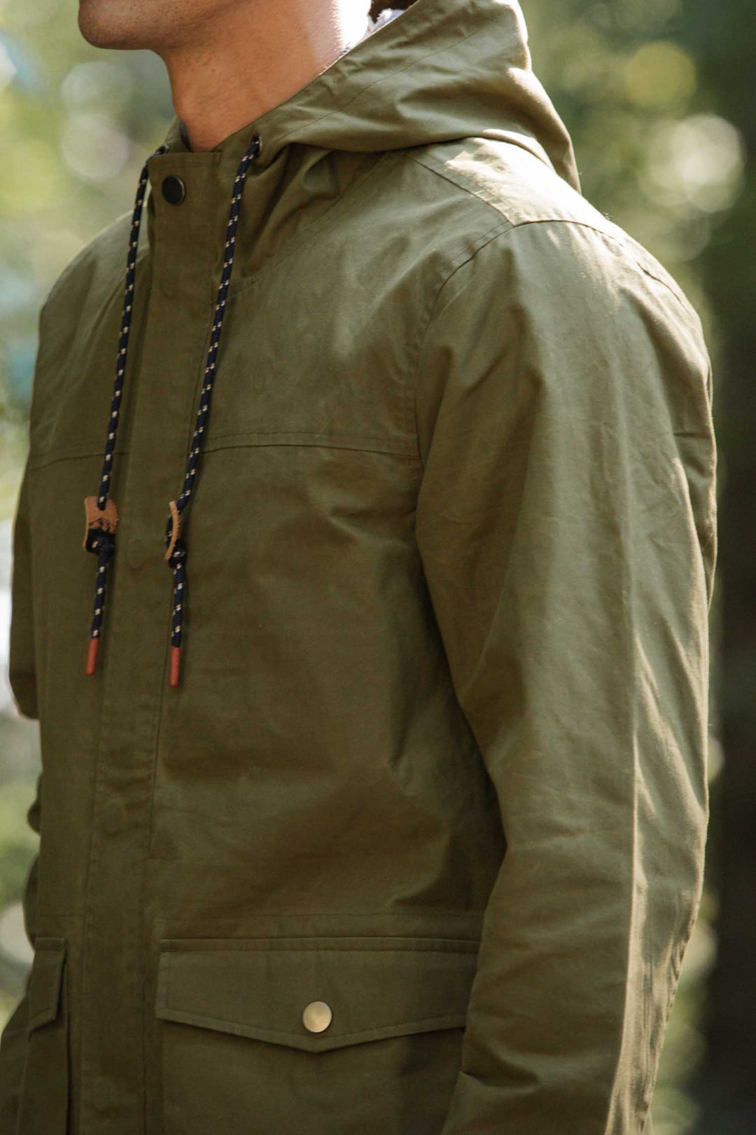 A person wearing the Bridge & Burn Marshall Jacket in olive waxed canvas, featuring black and white drawstrings and buttoned pockets, stands outdoors in soft natural light.